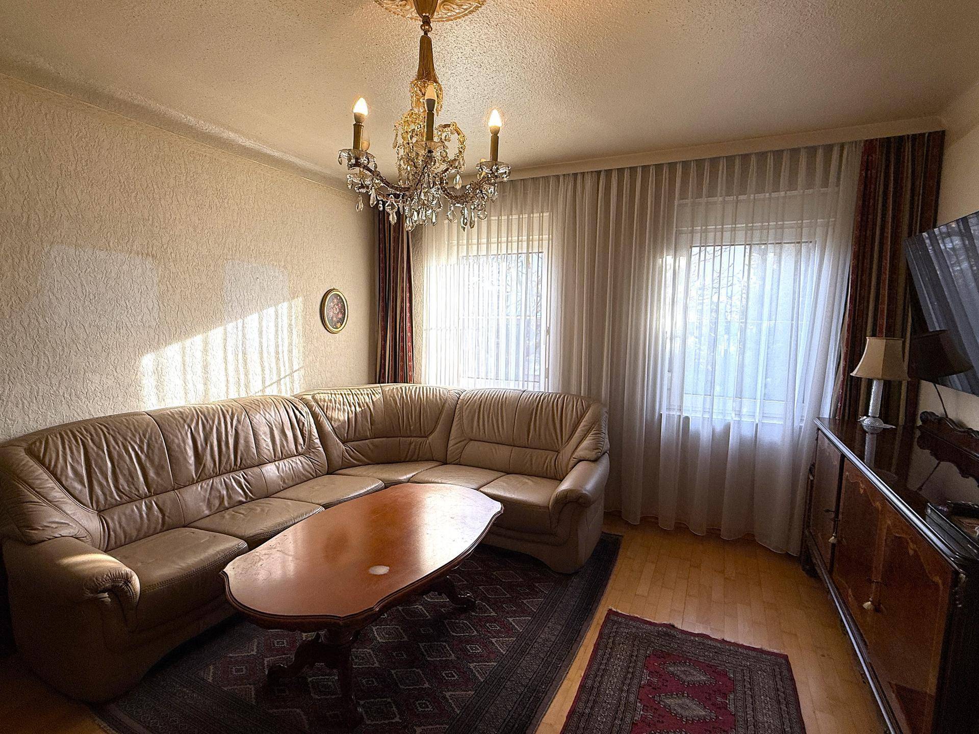 Wohnzimmer mit beiger Ledercouch, Kronleuchter und Teppichboden.