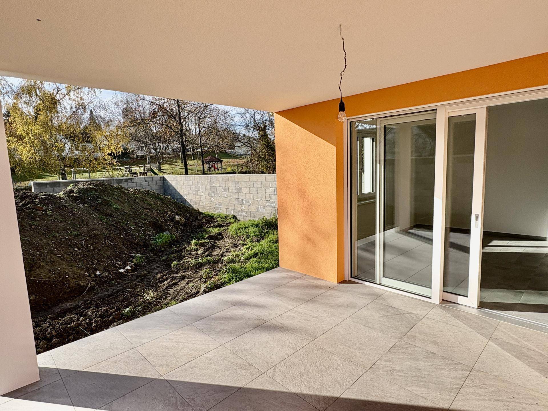 Überdachte Terrasse mit Fliesenboden und Blick auf den Garten und Spielplatz in der Ferne.