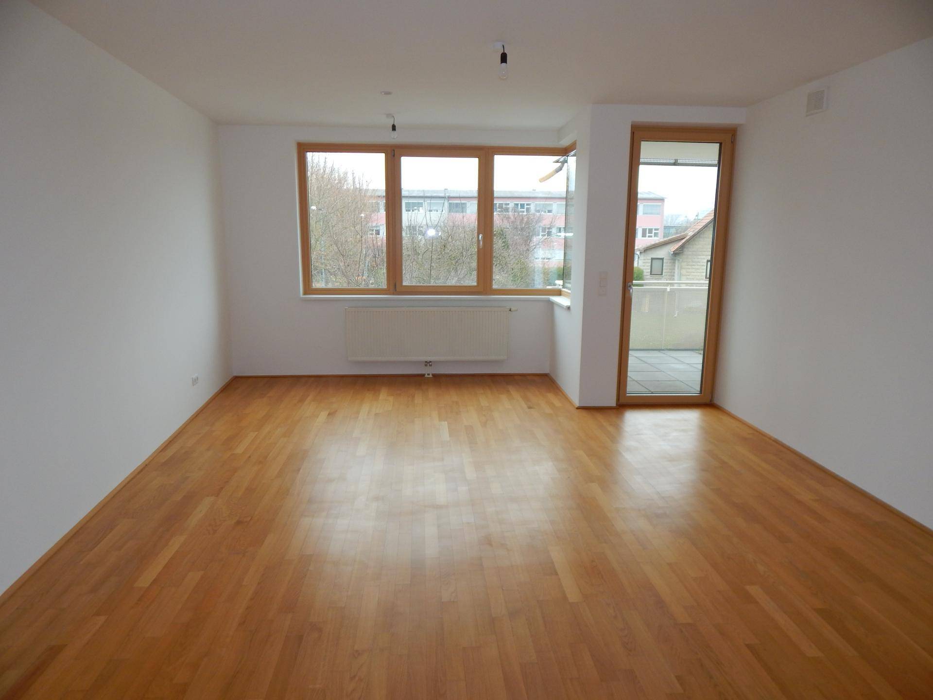 Helles, leeres Zimmer mit Holzboden, großen Fenstern und Zugang zu einem Balkon.
