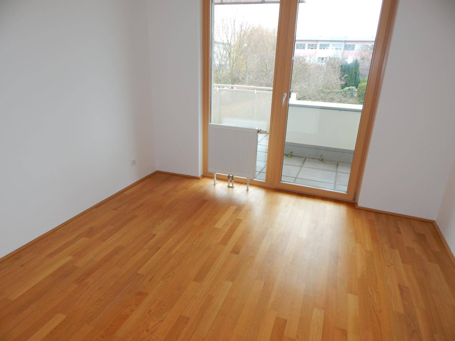 Leeres Zimmer mit Parkettboden und Balkonzugang, bietet viel natürliches Licht.