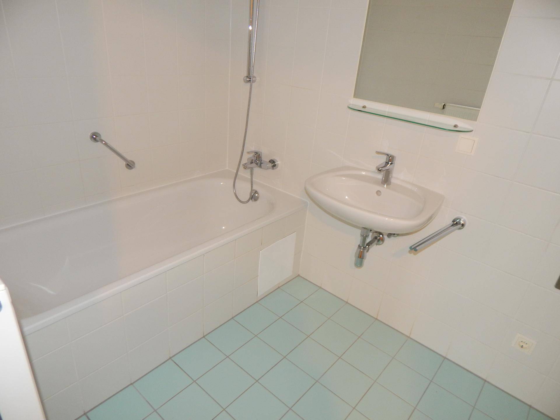 Badezimmer mit Badewanne, Waschbecken und hellblauen Fliesen, ausgestattet mit Haltegriffen.