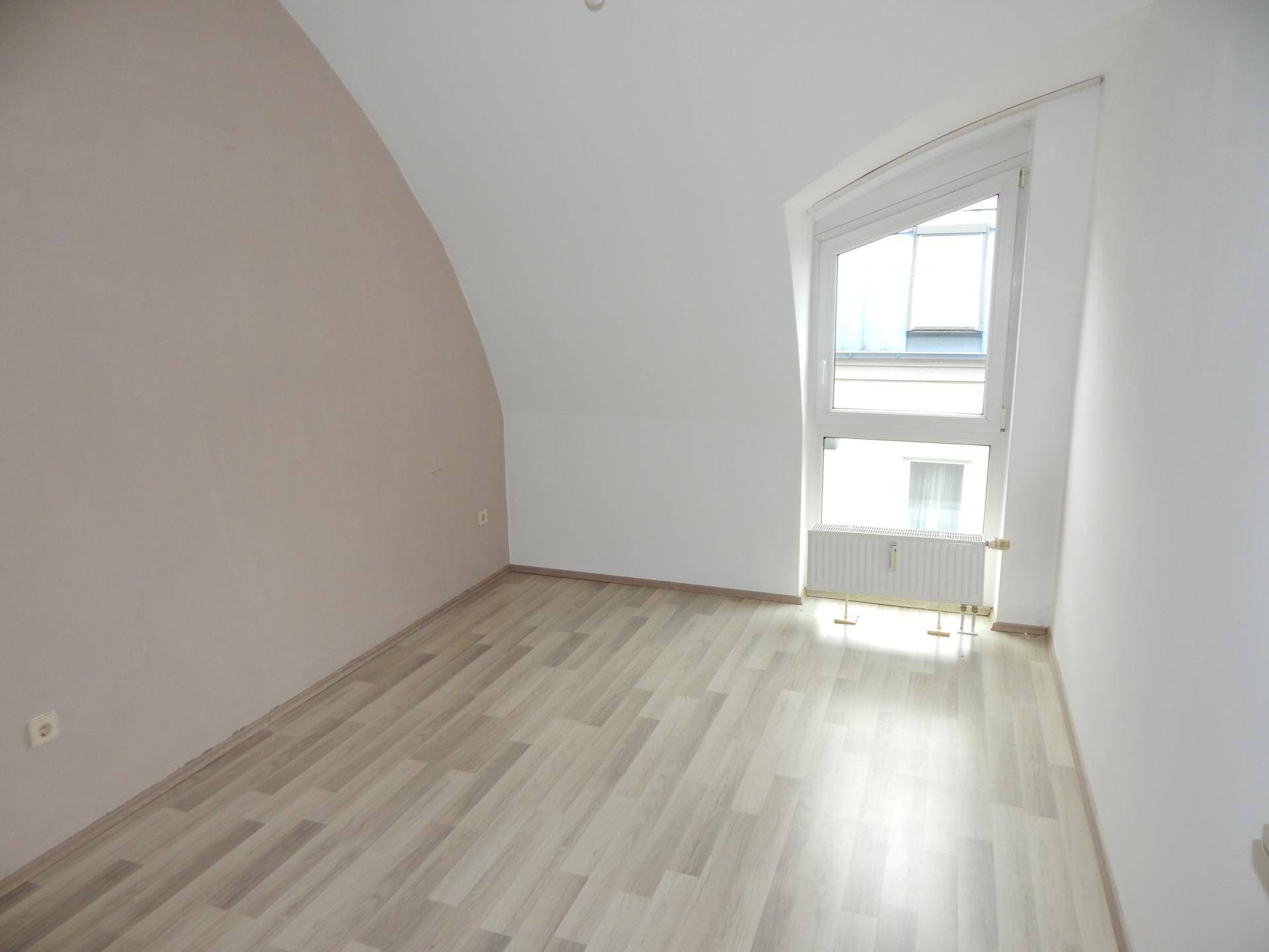 Helles Zimmer mit hellem Laminatboden, Dachschräge und einem großen Fenster.