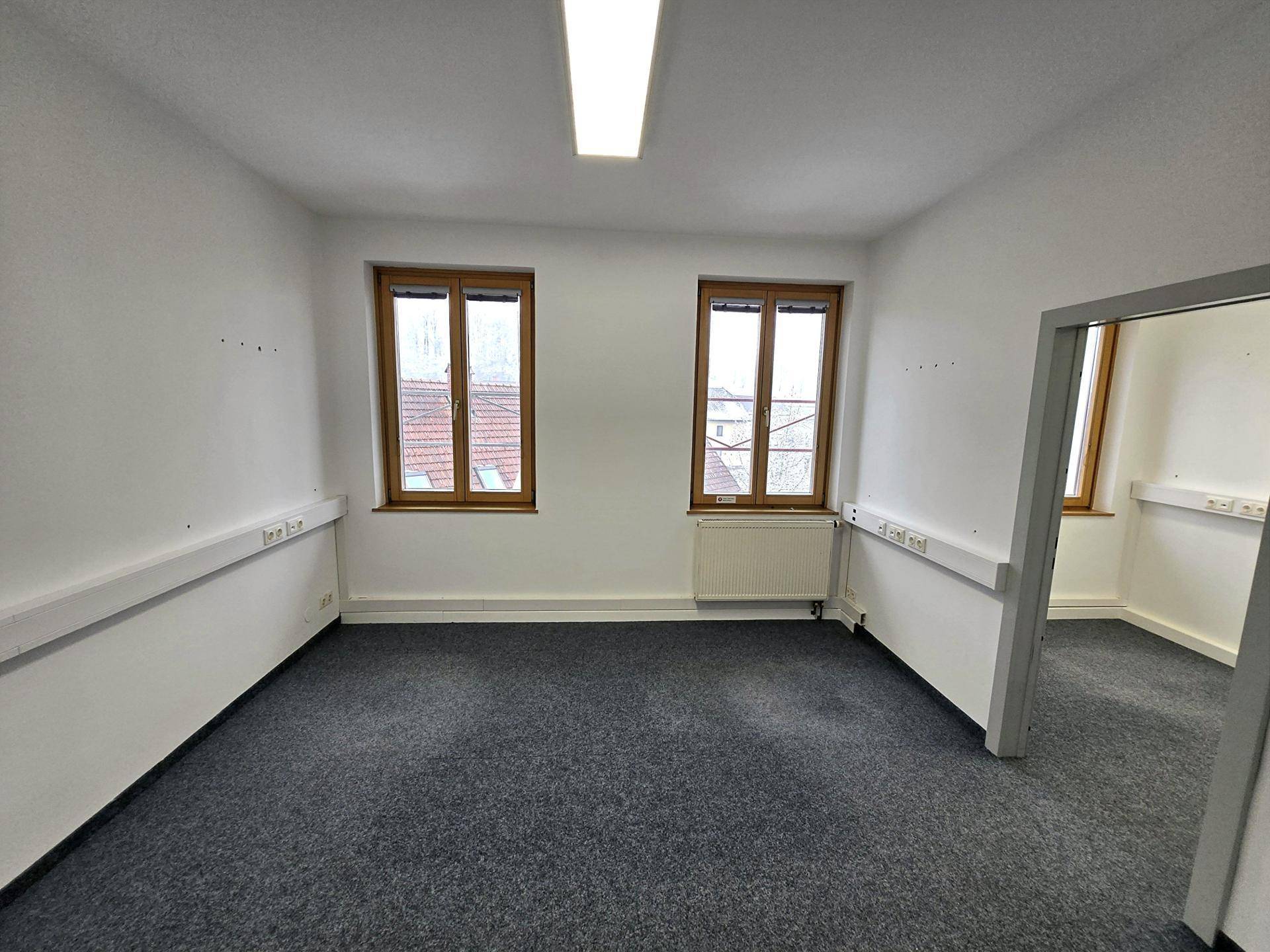 Geräumiger Büroraum mit Teppichboden, weißen Wänden und zwei Fenstern, die für eine helle Atmosphäre sorgen.