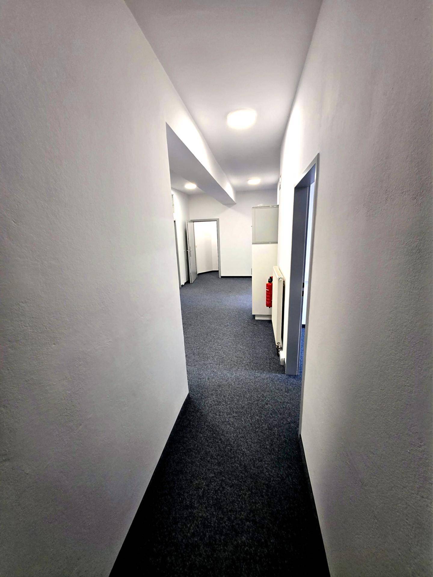 Langer, heller Flur mit Teppichboden und weißen Wänden, der zu verschiedenen Räumen und Türen führt.