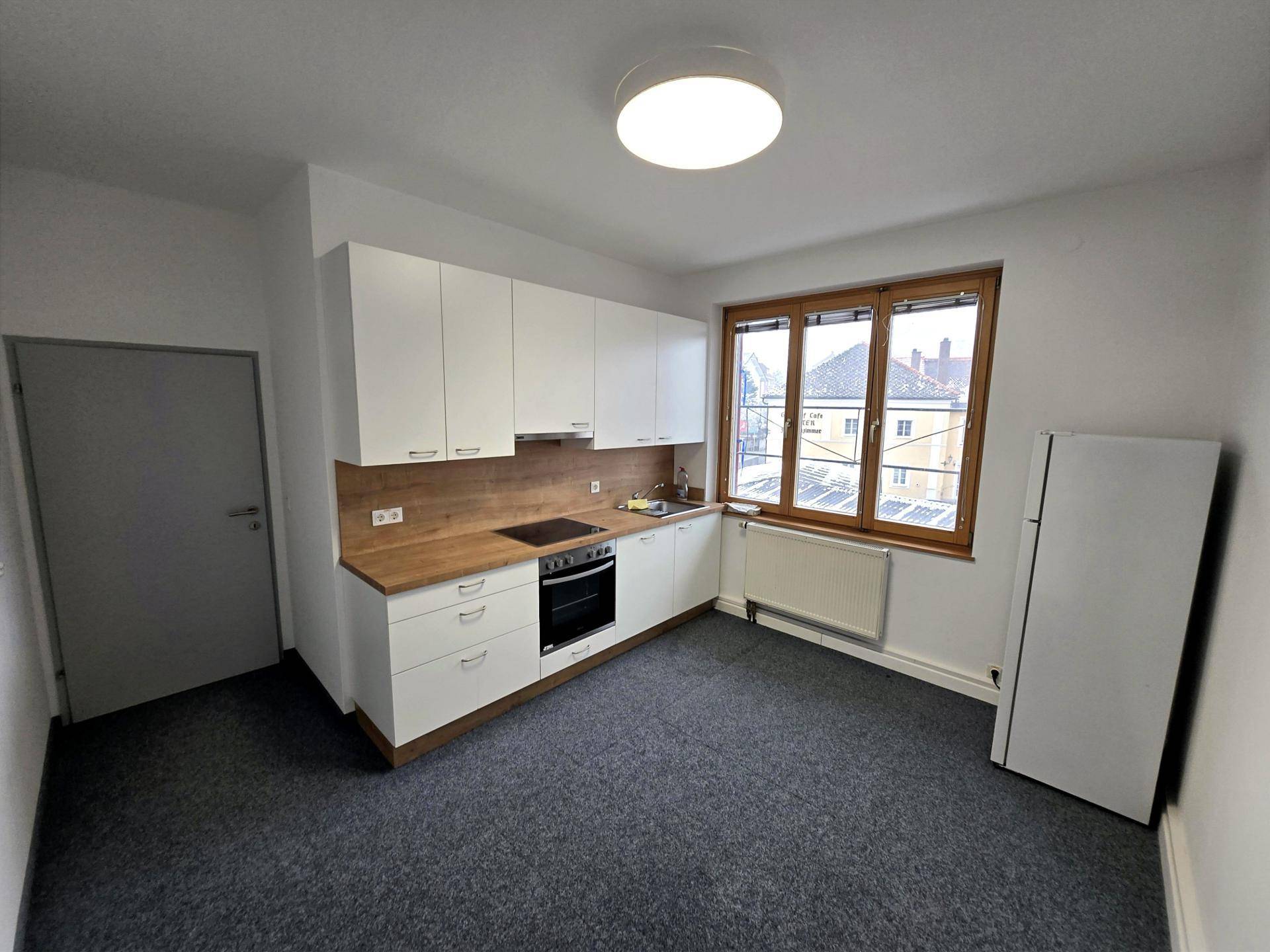 Kleine Küche mit weißen Schränken, Holzarbeitsplatte und Fenster in einem Raum mit Teppichboden.