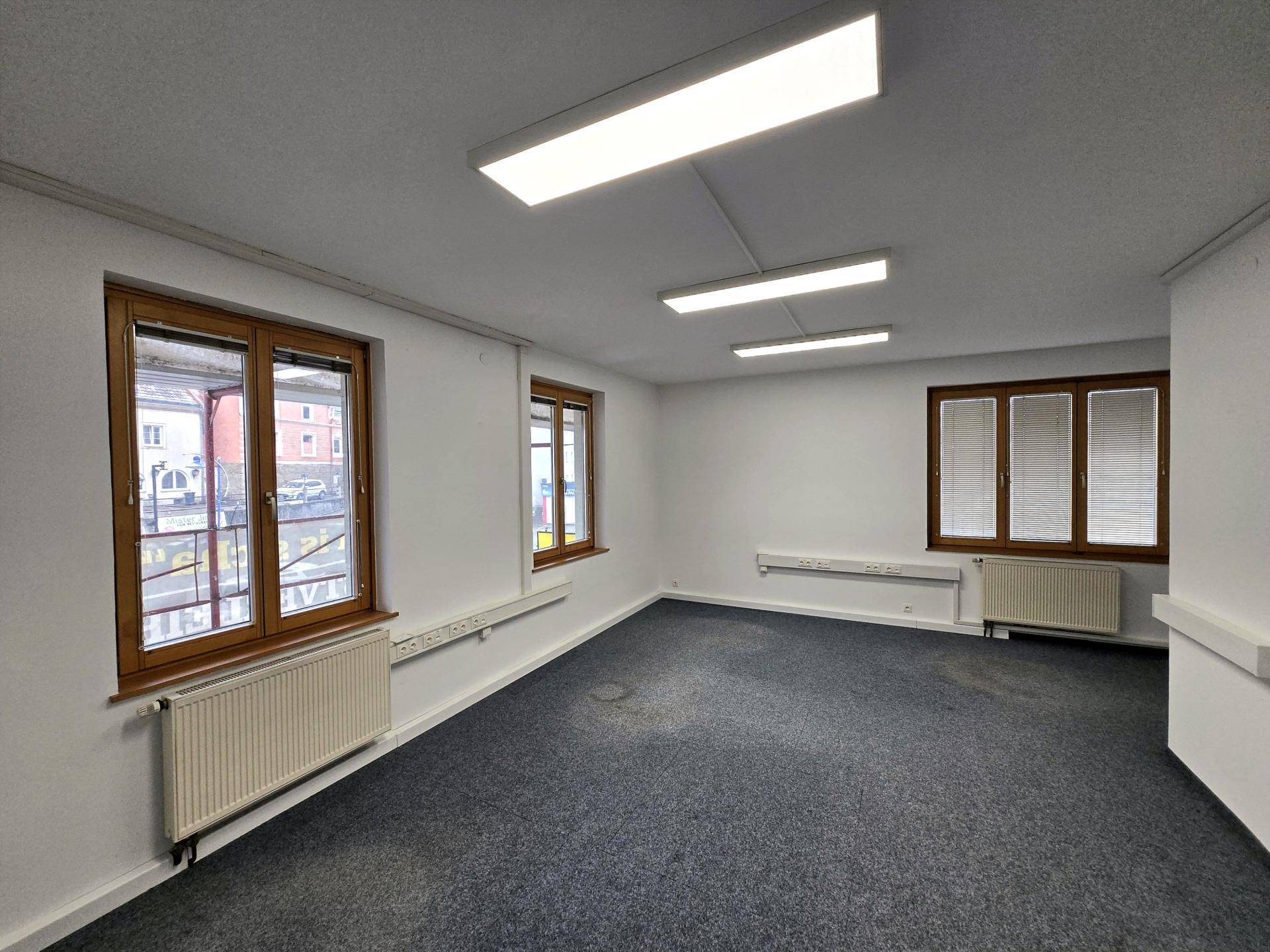 Leerer Büroraum mit Teppichboden, weißen Wänden und drei Fenstern, die viel Tageslicht hereinlassen.