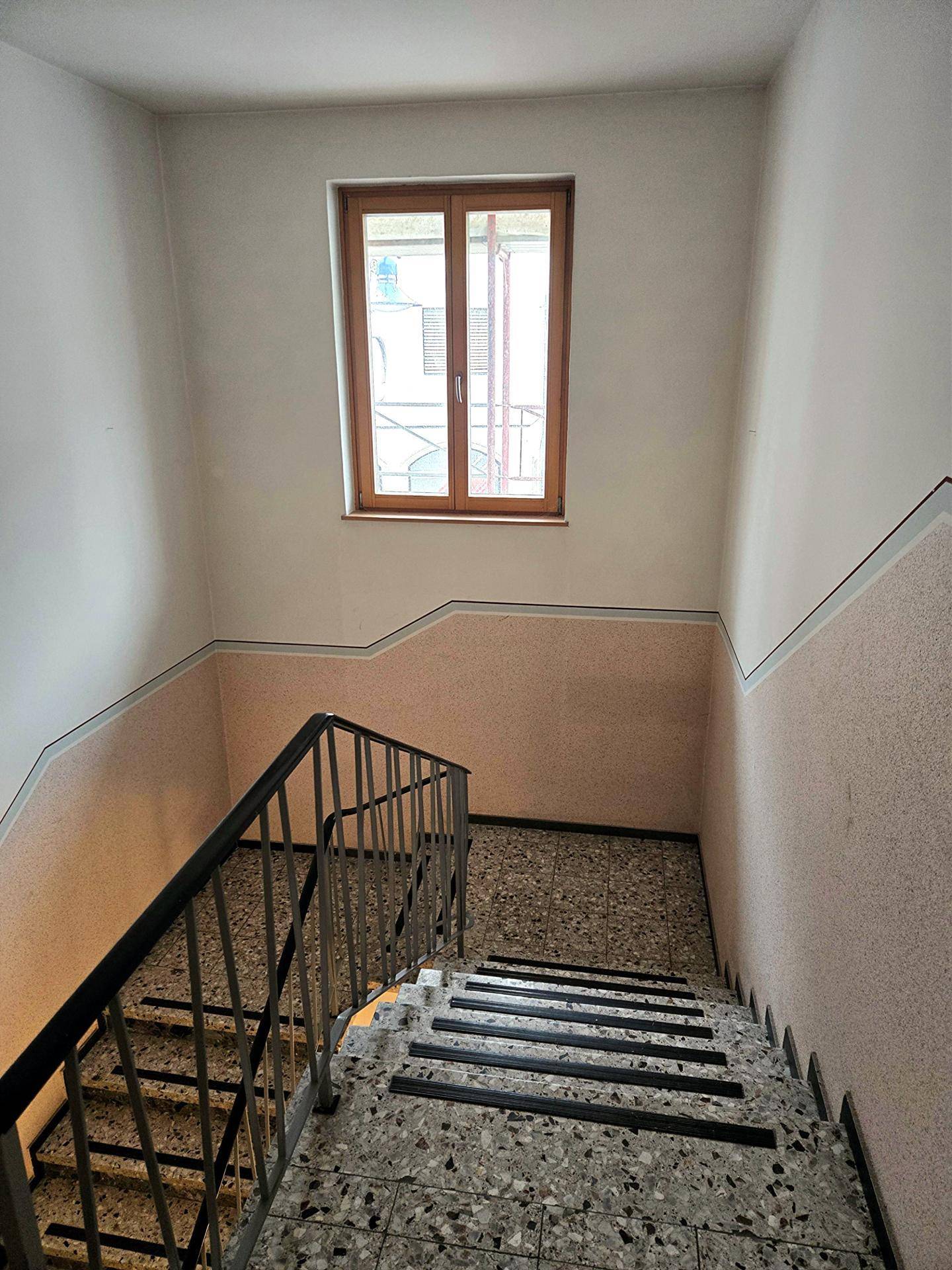 Treppenhaus mit Terrazzoboden, Metallgeländer und einem Fenster, das den Bereich mit Tageslicht versorgt.