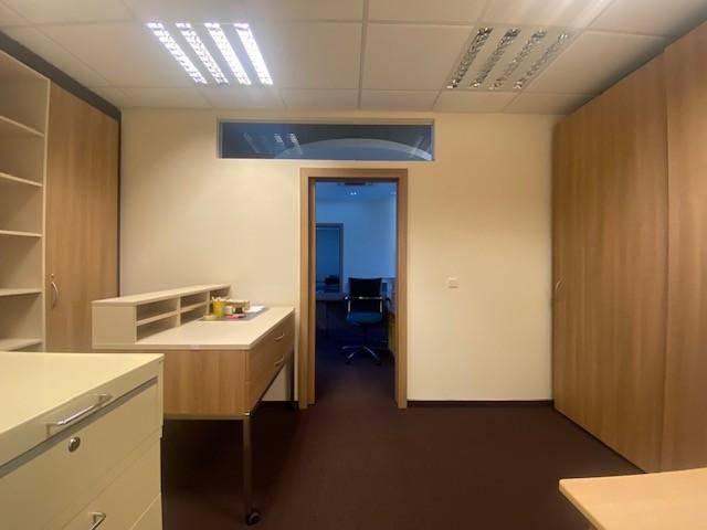 Büroraum mit Einbauschränken, einem Arbeitstisch und Teppichboden, der einen Blick in einen weiteren Raum bietet.