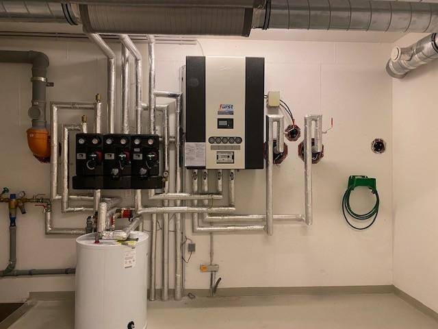 Technikraum im Keller mit Heizungsanlage, zahlreichen Rohren und einem Warmwasserspeicher.