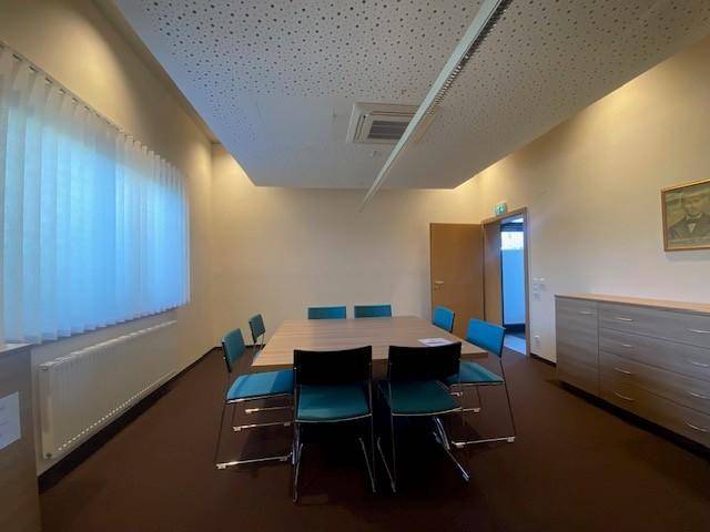 Geräumiger Konferenzraum mit großem Tisch, bequemen Stühlen und einem Fenster mit vertikalen Jalousien.