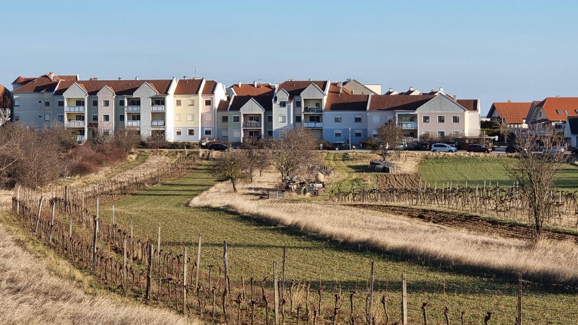 Mehrere Wohngebäude mit Balkonen und Blick auf Weinberge und Felder unter blauem Himmel.