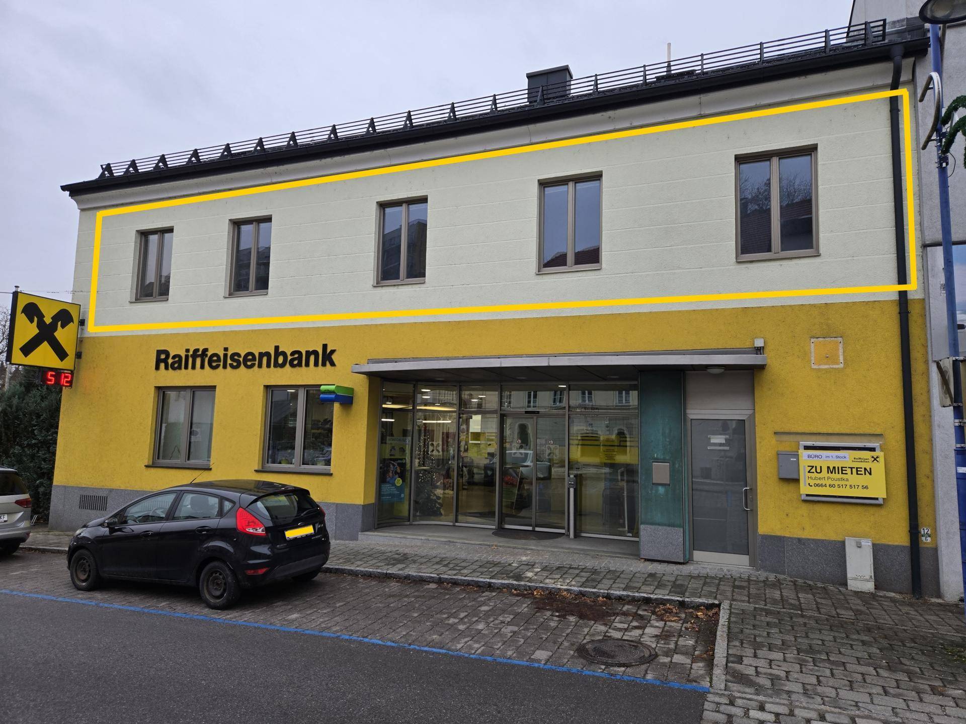Außenansicht eines Mehrzweckgebäudes mit gelber Bankfiliale im Erdgeschoss und einer zur Miete stehenden Obergeschosswohnung.