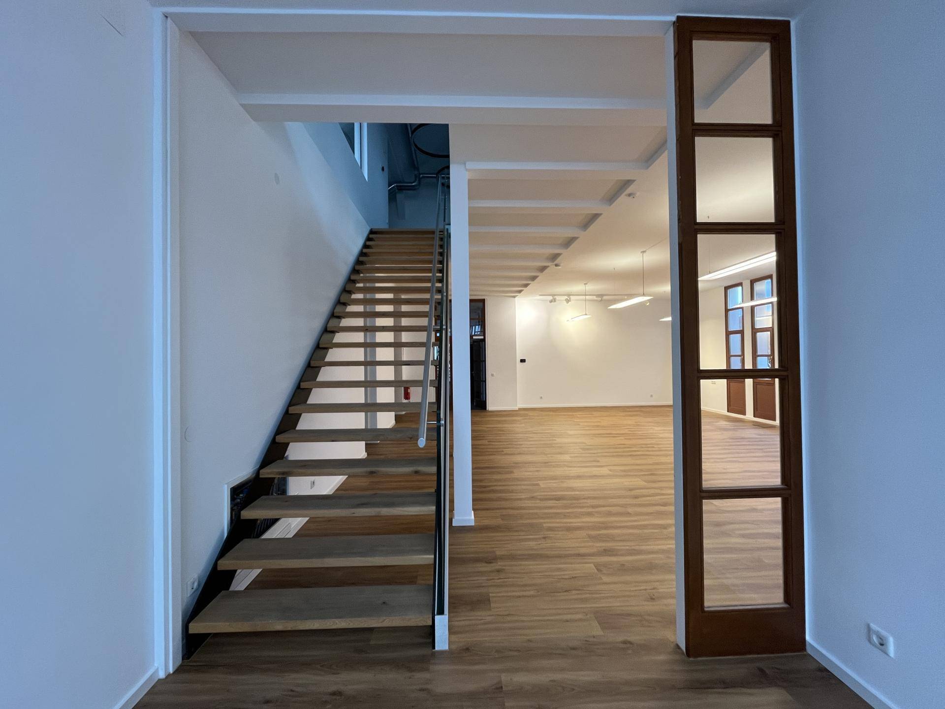 Offener Innenraum mit Holztreppe, Holzboden und Blick in einen großen, hellen Bereich.