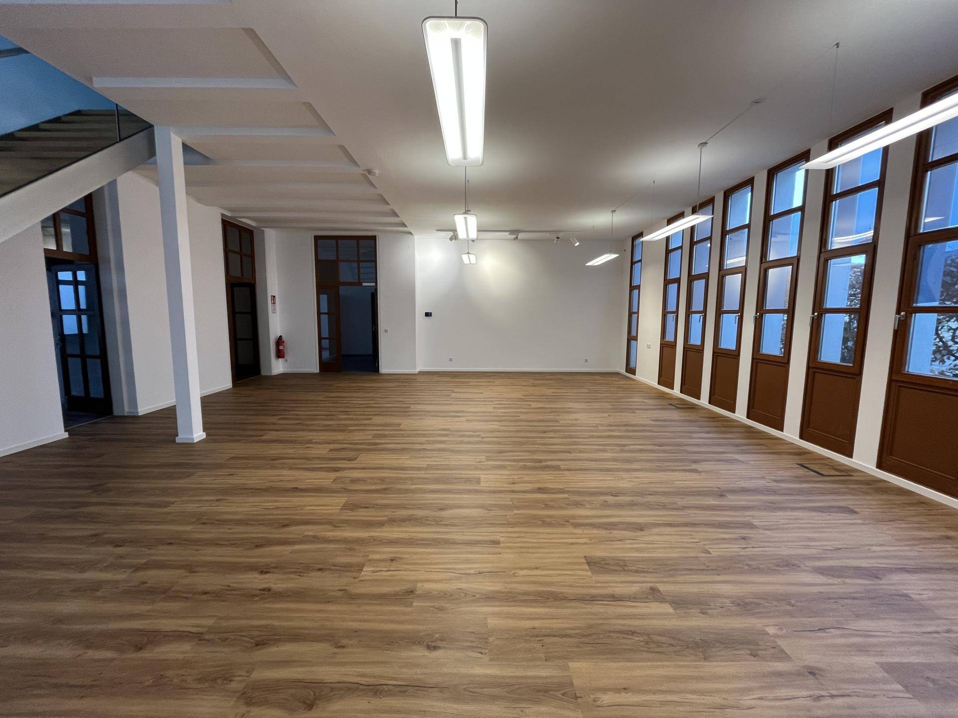 Großer, heller Raum mit Holzboden, vielen Fenstern und moderner Deckenbeleuchtung.