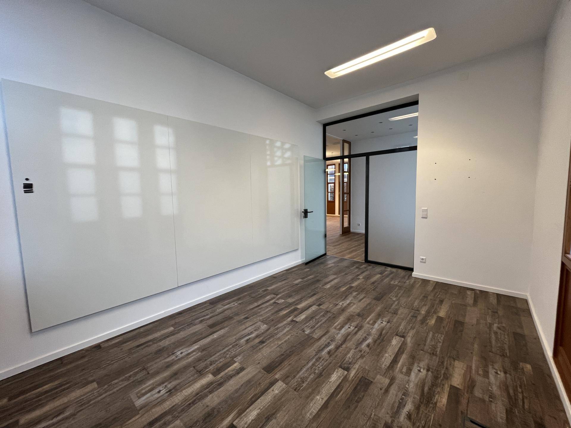 Büro mit Holzboden, großer Whiteboard-Wand und Glastür zu einem weiteren Raum.
