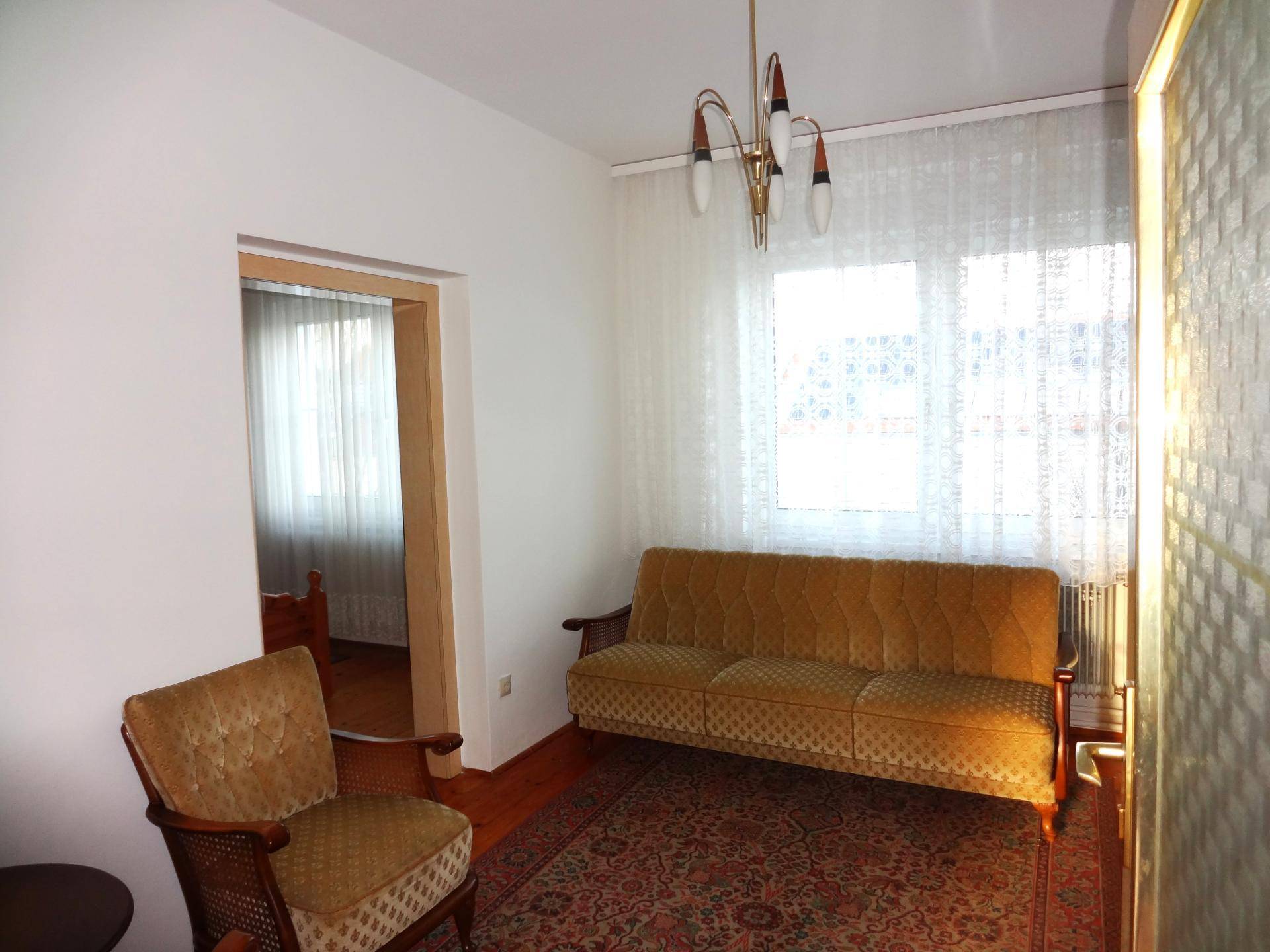 Gemütliches Zimmer mit gelbem Sofa, Sessel und einem Fenster mit Vorhängen.