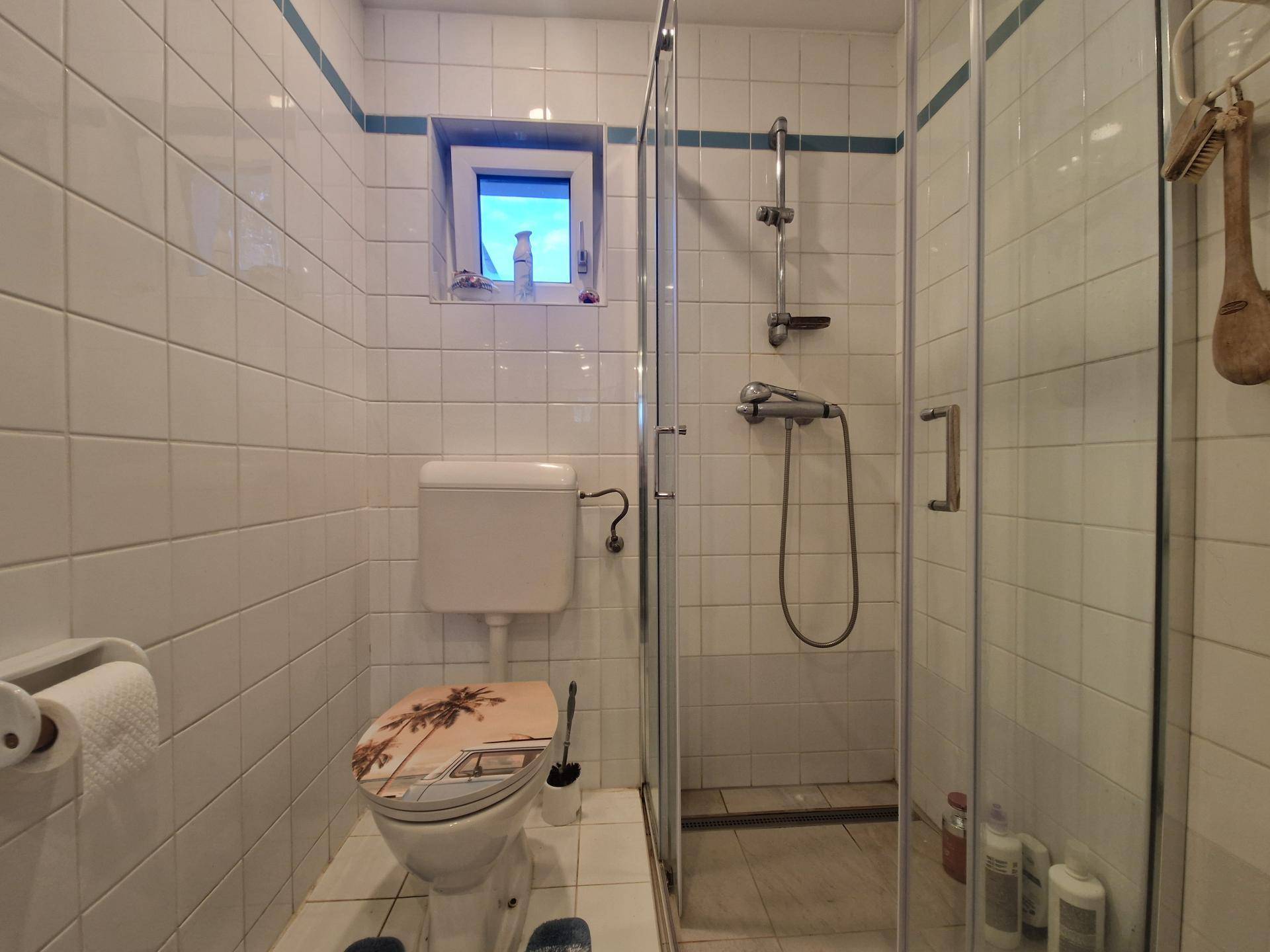 Kleines Badezimmer mit weißen Fliesen, Duschkabine und Fenster für Tageslicht.