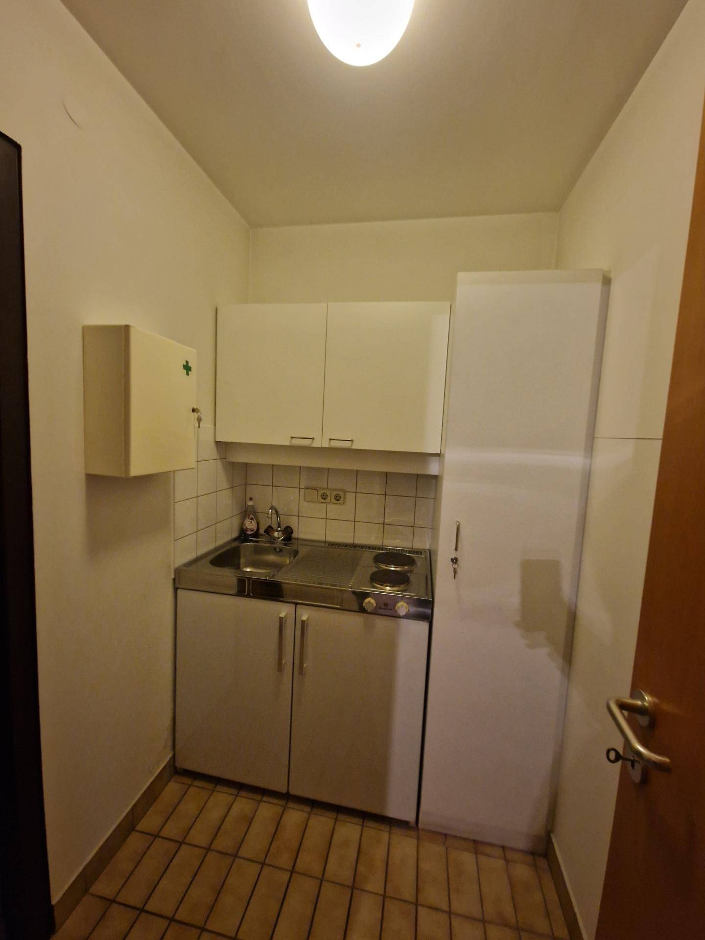 Kleine Küchenzeile mit weißen Schränken, Spüle, Kochfeld und Kühlschrank.