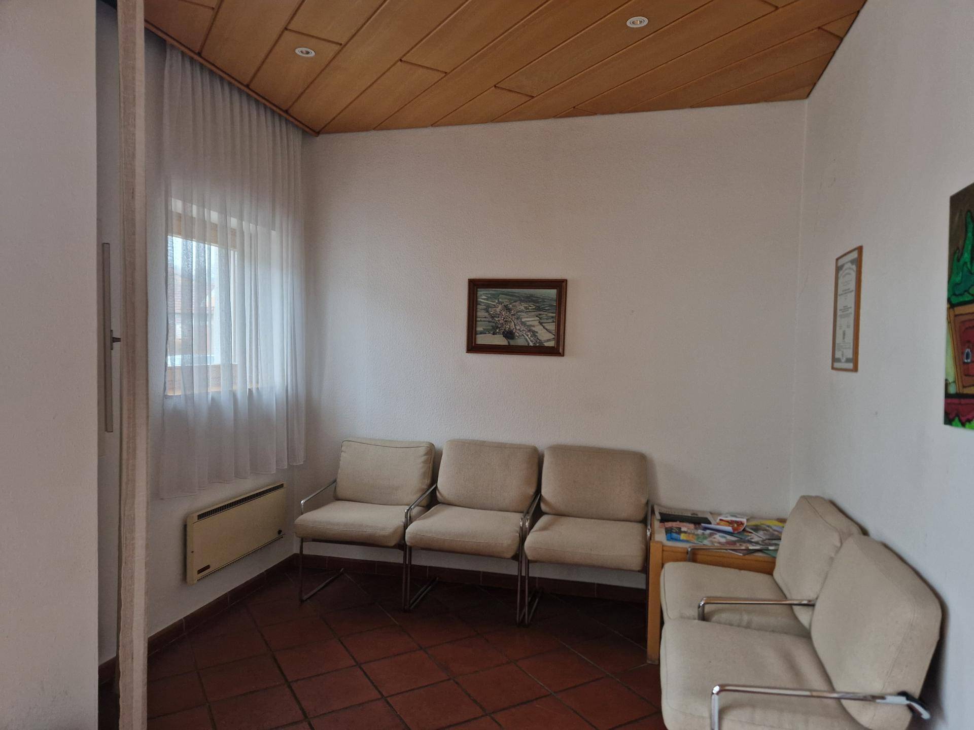 Wartebereich mit Terrakottafliesen, bequemen Stühlen und einem Fenster mit Vorhängen.