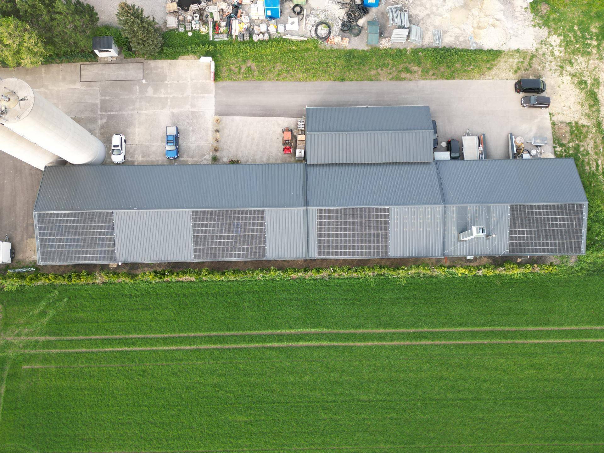 Luftaufnahme eines großen Industriegebäudes mit zahlreichen Solarmodulen auf dem grauen Metalldach, umgeben von Grünflächen und einem Hof.