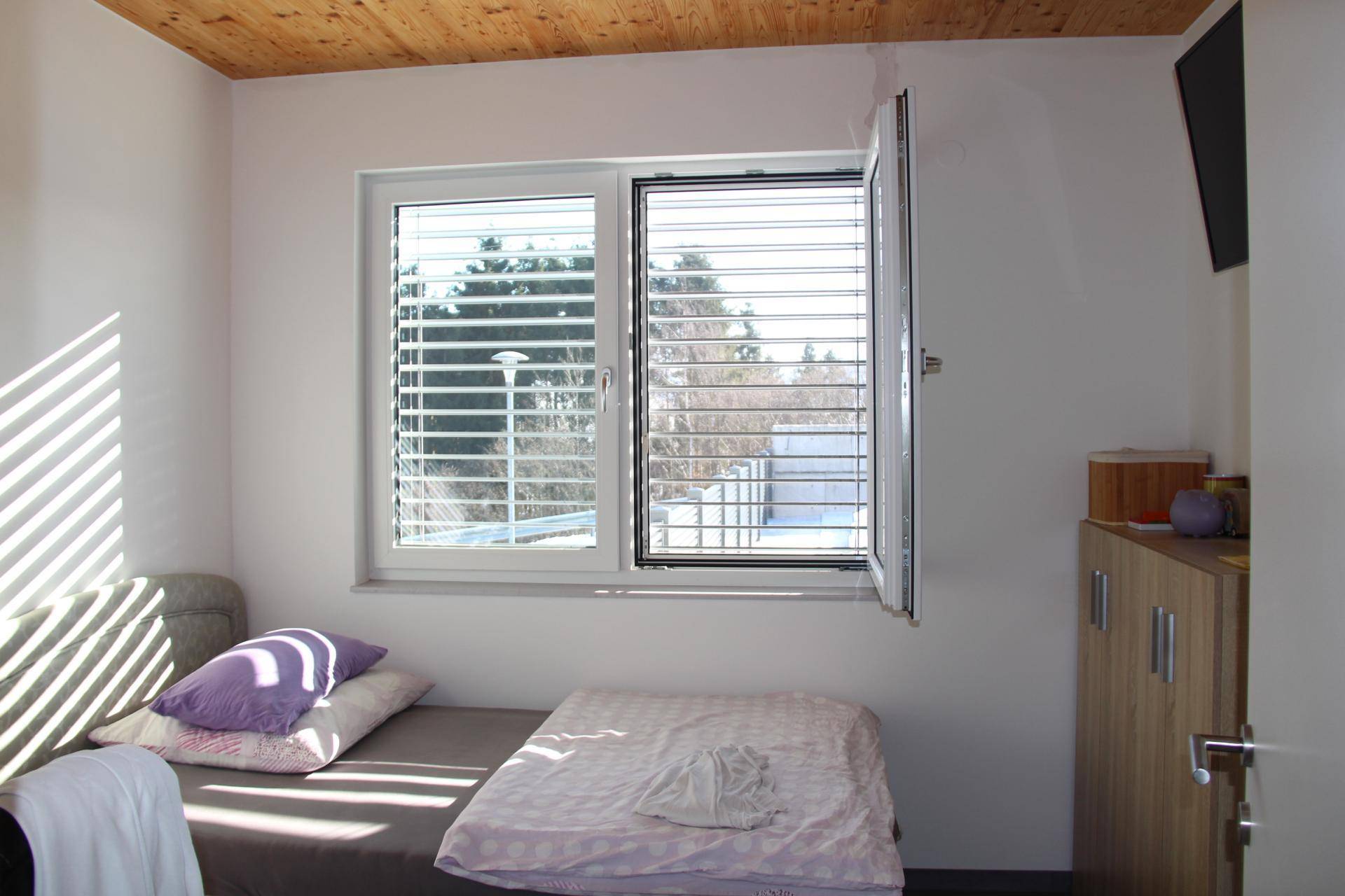 Helles Schlafzimmer mit Holzbalkendecke, Laminatboden und Fenster mit Jalousien.