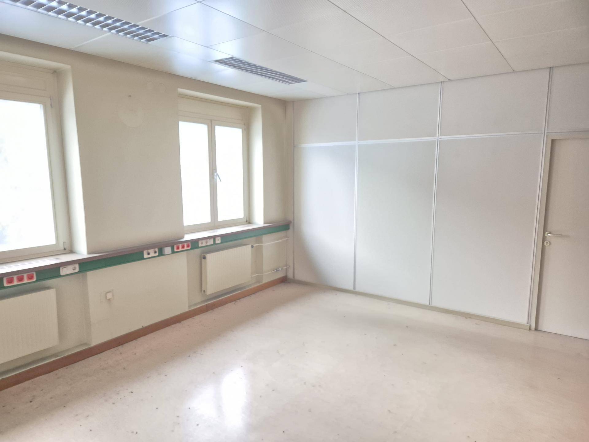 Heller, leerer Raum mit mehreren Fenstern, Heizkörpern und einer Reihe von Steckdosen entlang der Wand.