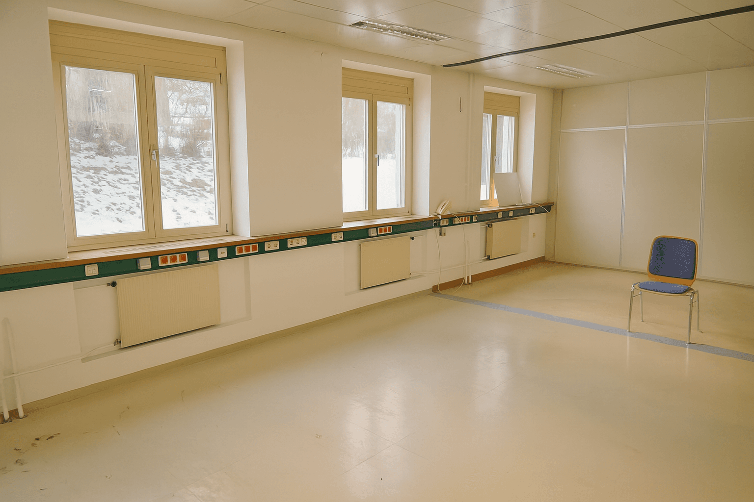 Geräumiger, leerer Raum mit mehreren Fenstern, Heizkörpern und einer Reihe von Steckdosen entlang der Wand.