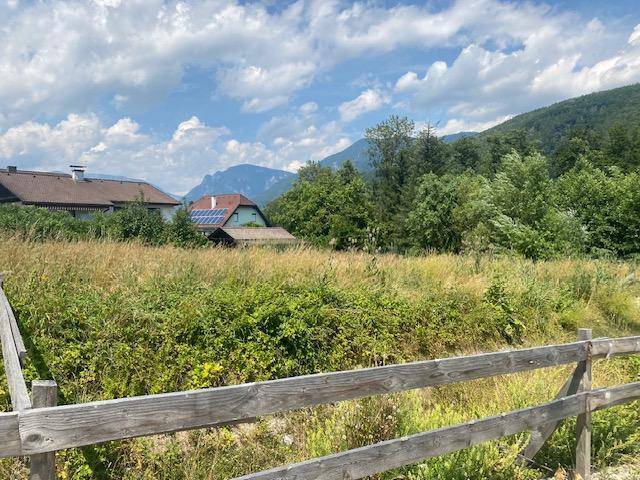 Blick über ein Feld mit Holzzaun auf traditionelle Häuser und bewaldete Berge im Hintergrund.