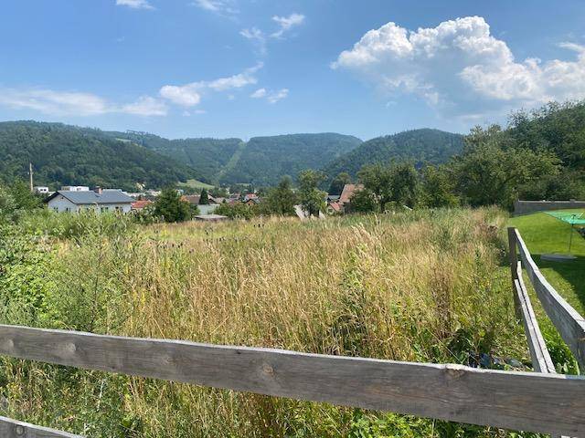 Weitläufige Landschaft mit Feldern, Holzzaun und Blick auf eine Siedlung vor bewaldeten Hügeln.