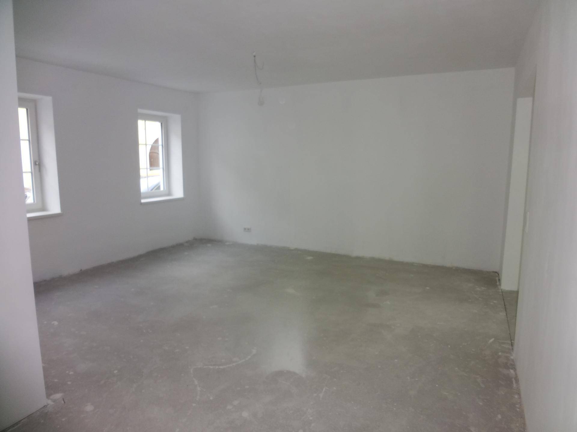 Geräumiger, unfertiger Raum mit zwei Fenstern und Betonboden, bereit für den Ausbau.