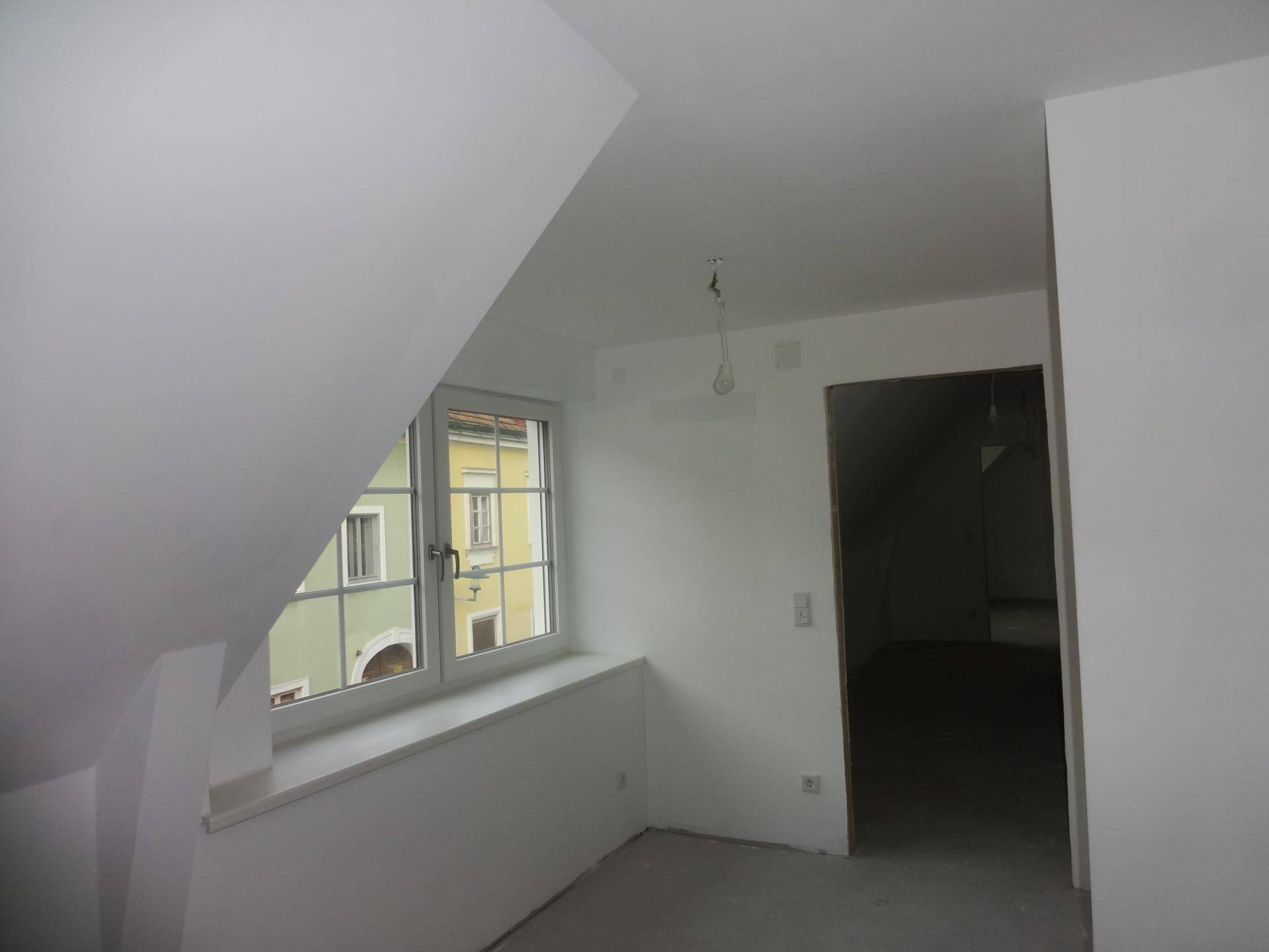 Unfertiger Dachgeschossraum mit Schrägdach, Fenster und Blick auf die Umgebung.
