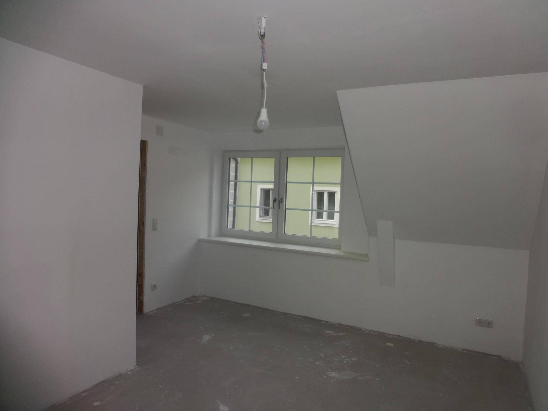 Unfertiger Dachgeschossraum mit Schrägdach, Fenster und Blick auf ein Nachbargebäude.