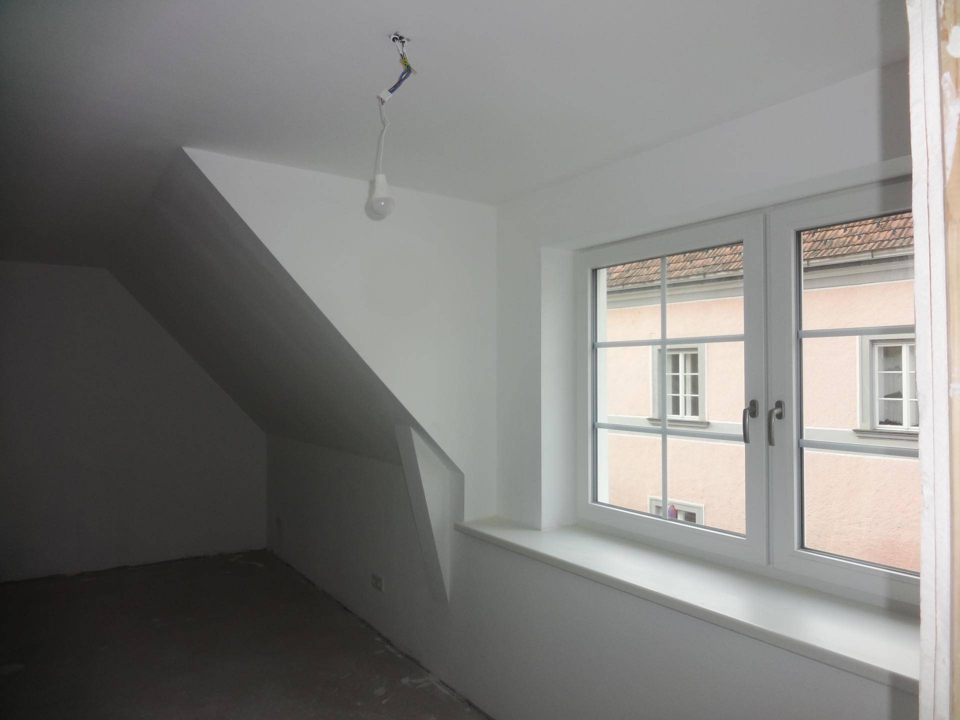 Unfertiger Dachgeschossraum mit Schrägdach, zwei Fenstern und Blick auf die Umgebung.
