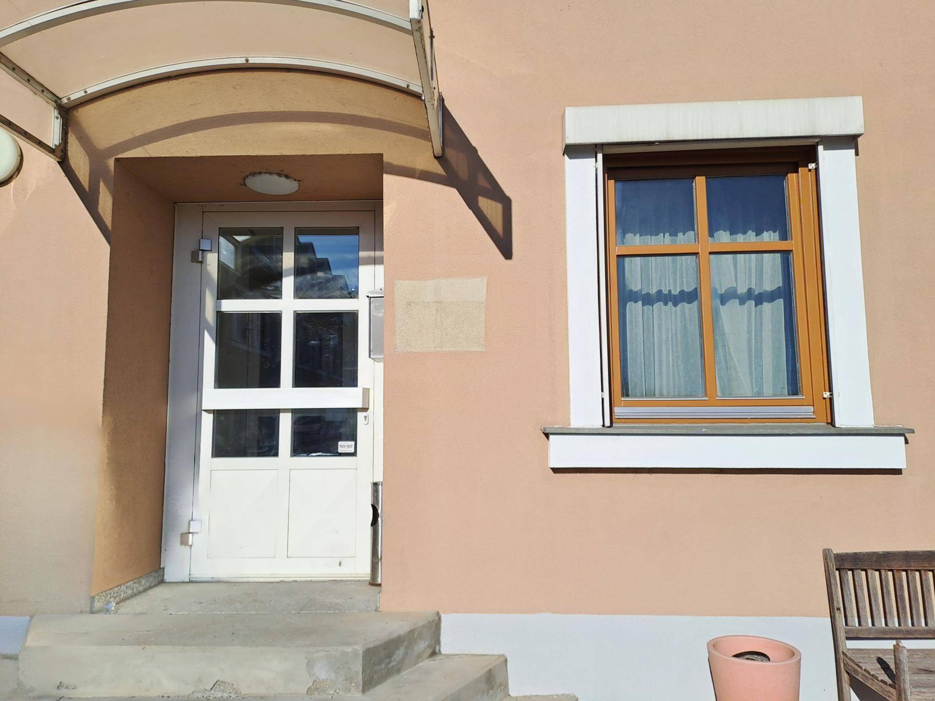 Eingangsbereich des Gebäudes mit Vordach, weißer Tür und Fenster mit Rollladen.