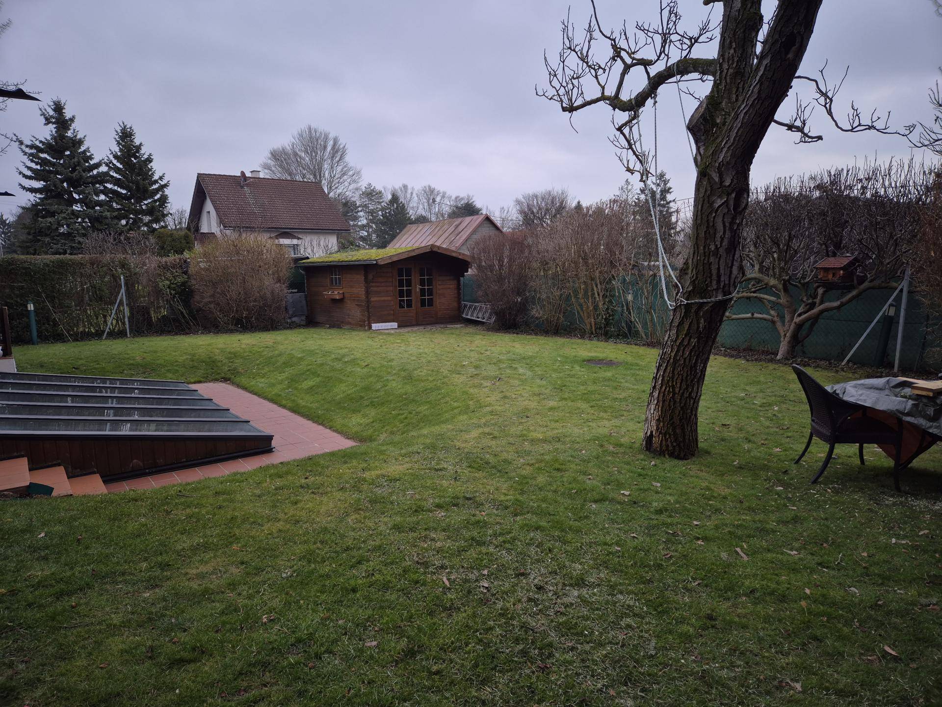 Weitläufiger Garten mit Rasenfläche, Gartenhaus und altem Baumbestand unter bewölktem Himmel.
