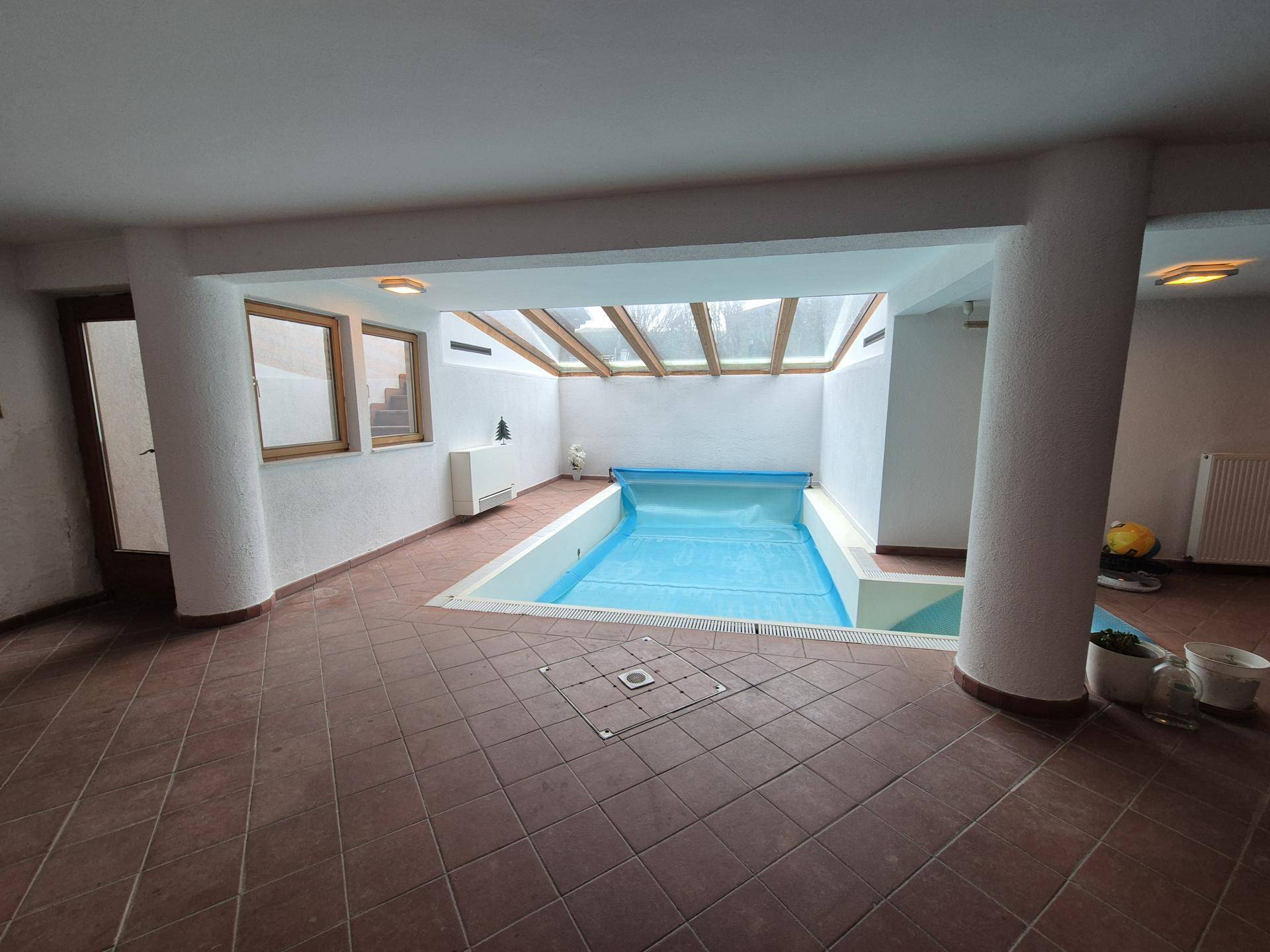Innenpoolbereich mit Tageslicht durch Glasdach und gefliestem Boden, umgeben von weißen Säulen.