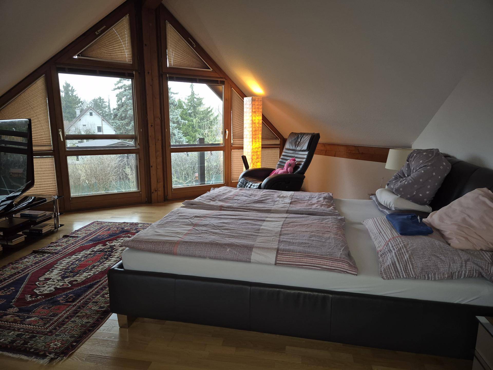 Schlafzimmer im Dachgeschoss mit schrägen Wänden, Holzbalken und großen Fenstern.