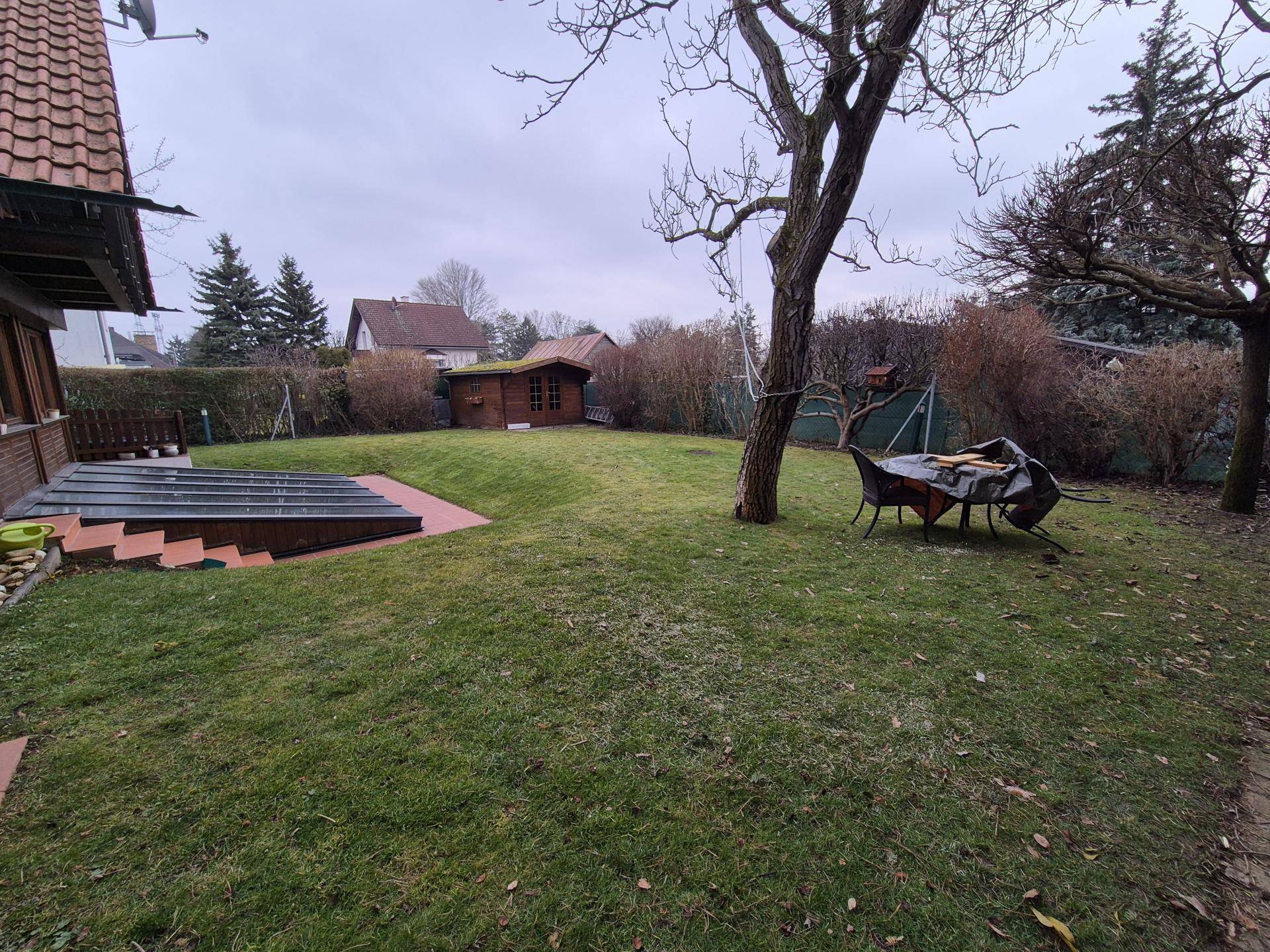 Weitläufiger Garten mit Rasenfläche, Gartenhaus und altem Baumbestand unter bewölktem Himmel.