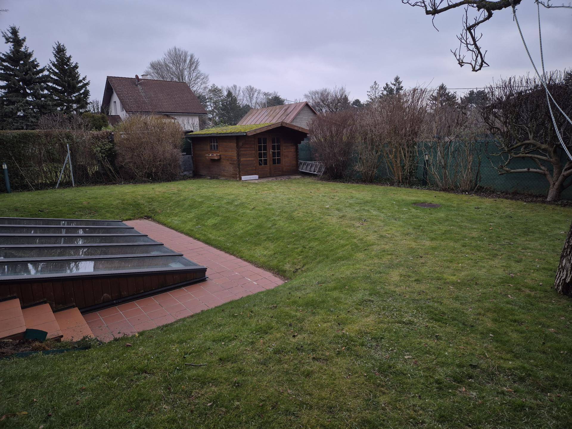 Weitläufiger Garten mit Rasenfläche, Gartenhaus und altem Baumbestand unter bewölktem Himmel.