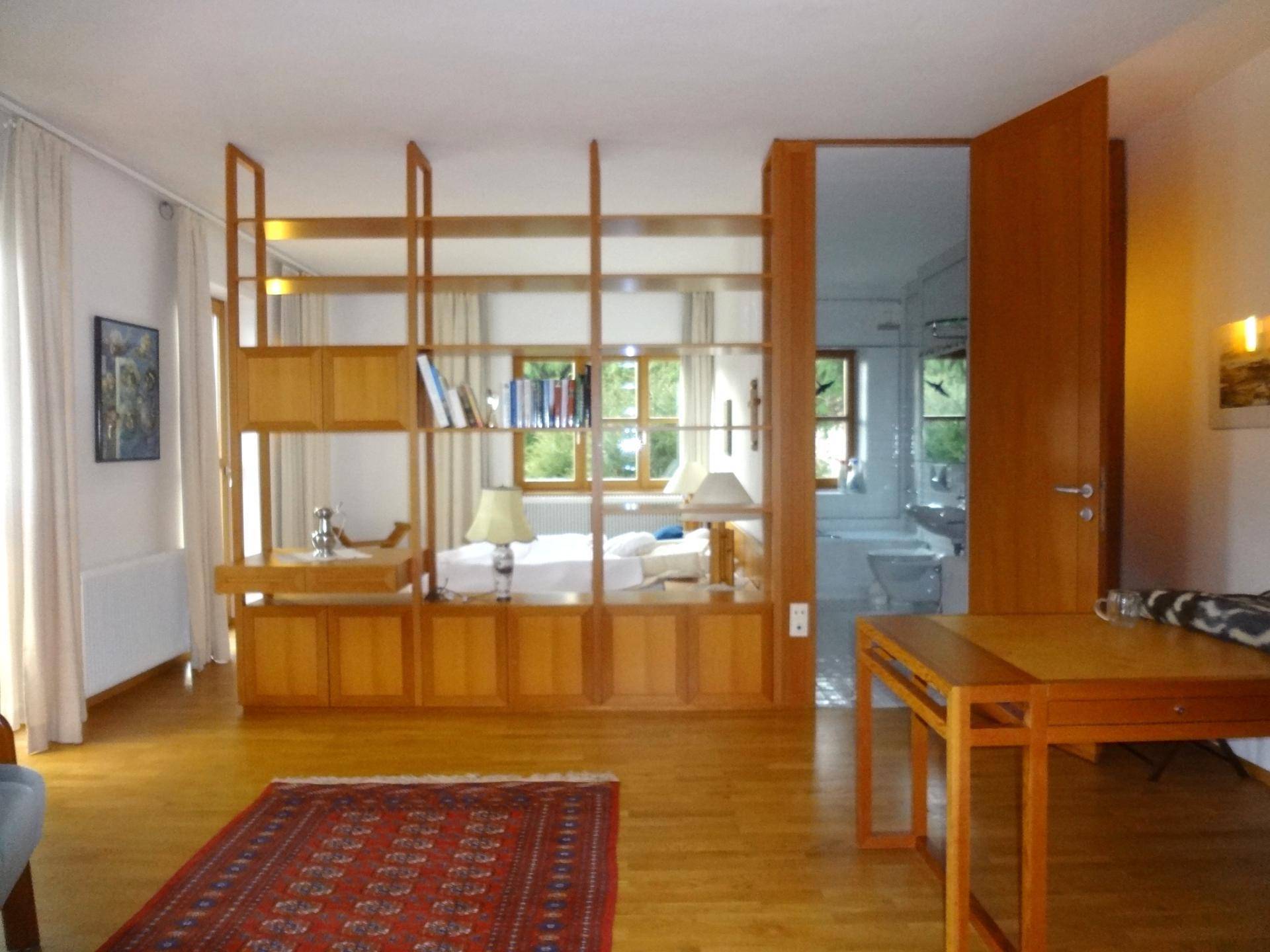 Helles Schlafzimmer mit Holzfußboden, einem großen Regal als Raumteiler und Blick ins Grüne.