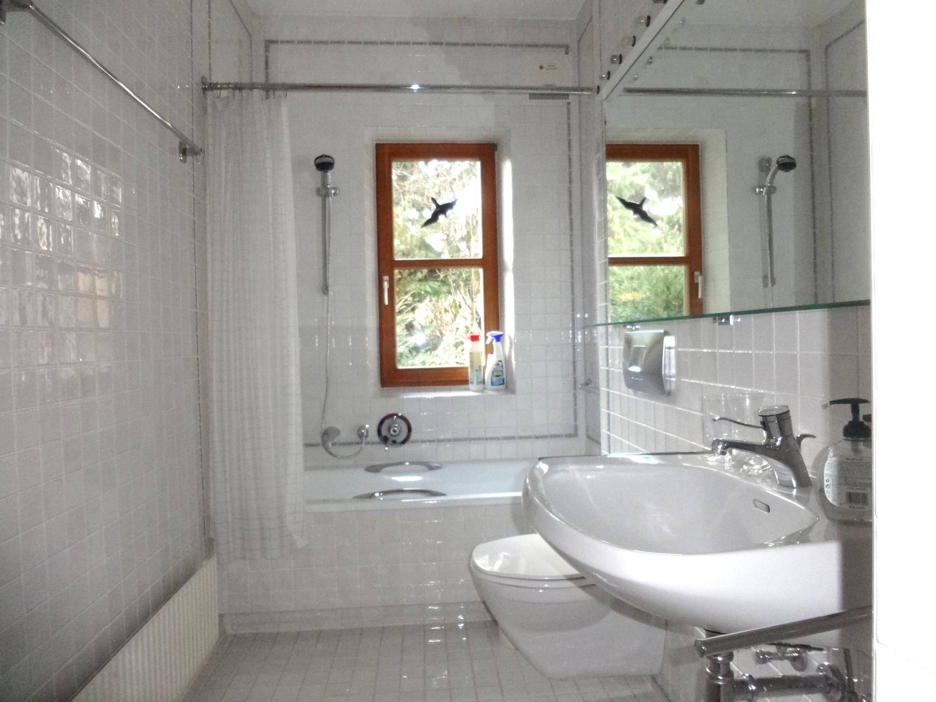 Badezimmer mit weißen Fliesen, einer Badewanne mit Duschfunktion und einem Fenster für Tageslicht.