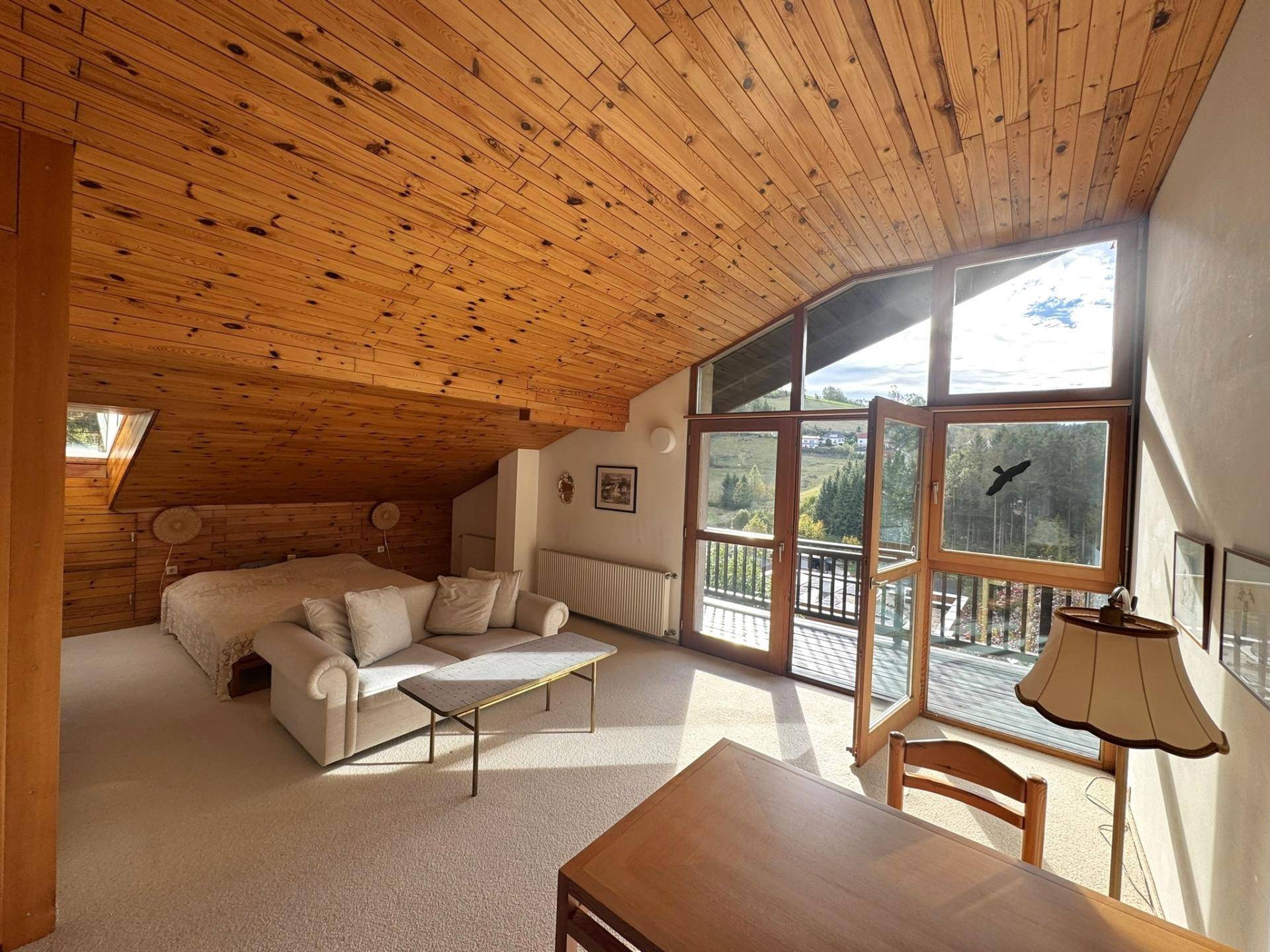 Helles Schlafzimmer im Dachgeschoss mit schräger Holzdecke, großen Fenstern und Balkonzugang.