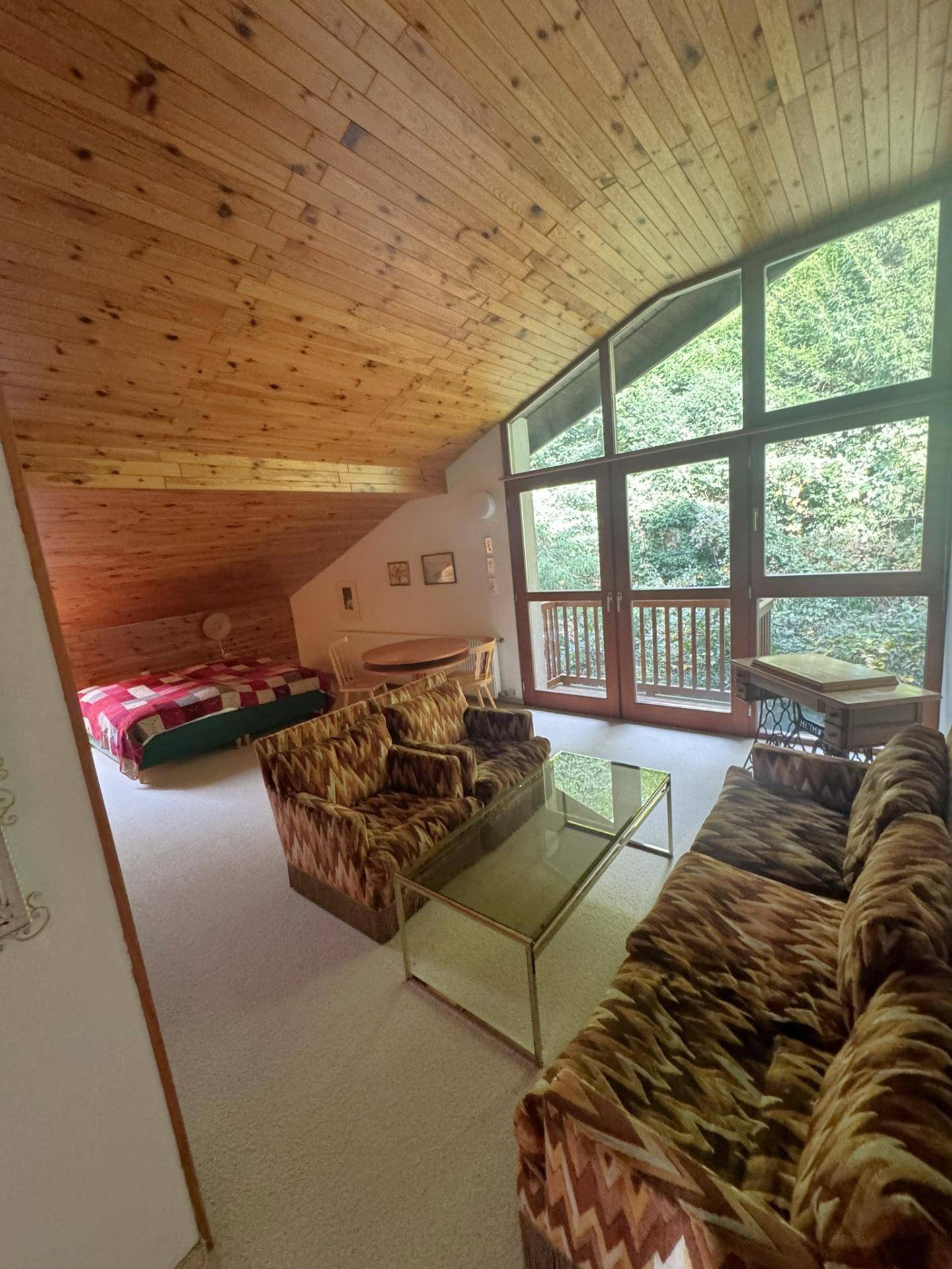 Geräumiger Wohnbereich im Dachgeschoss mit schräger Holzdecke und großen Fenstern mit Waldblick.