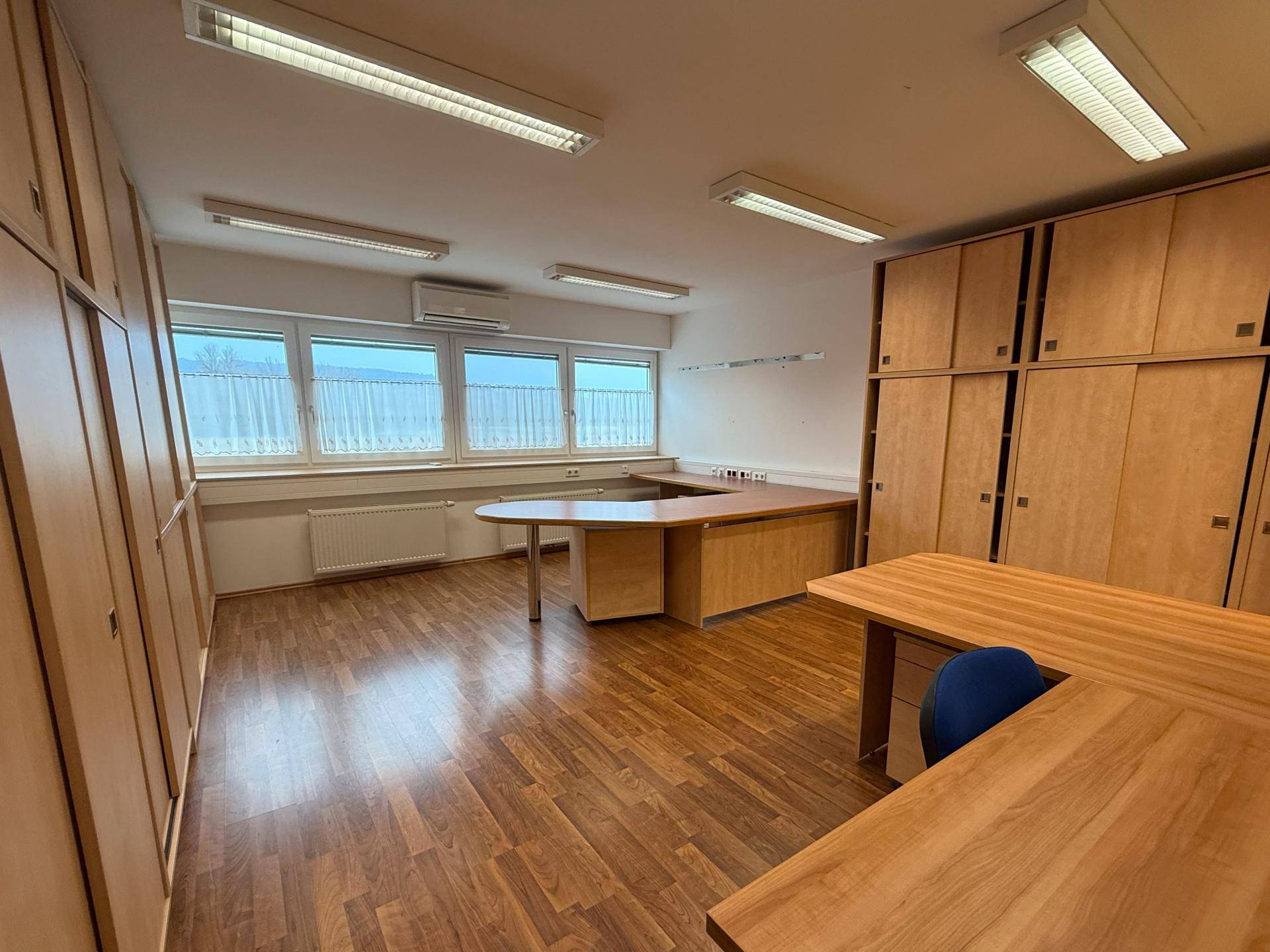 Helles Büro mit großen Fenstern, Klimaanlage und umfangreichen Holzschränken, ideal für konzentriertes Arbeiten.