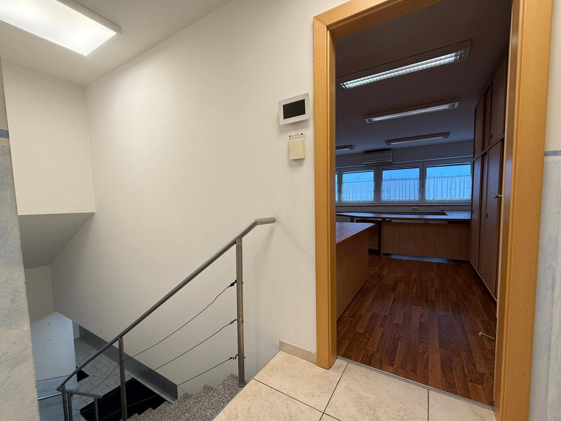 Treppenaufgang mit hellem Fliesenboden und Blick in ein angrenzendes Büro mit Holzmöbeln, gut beleuchtet.