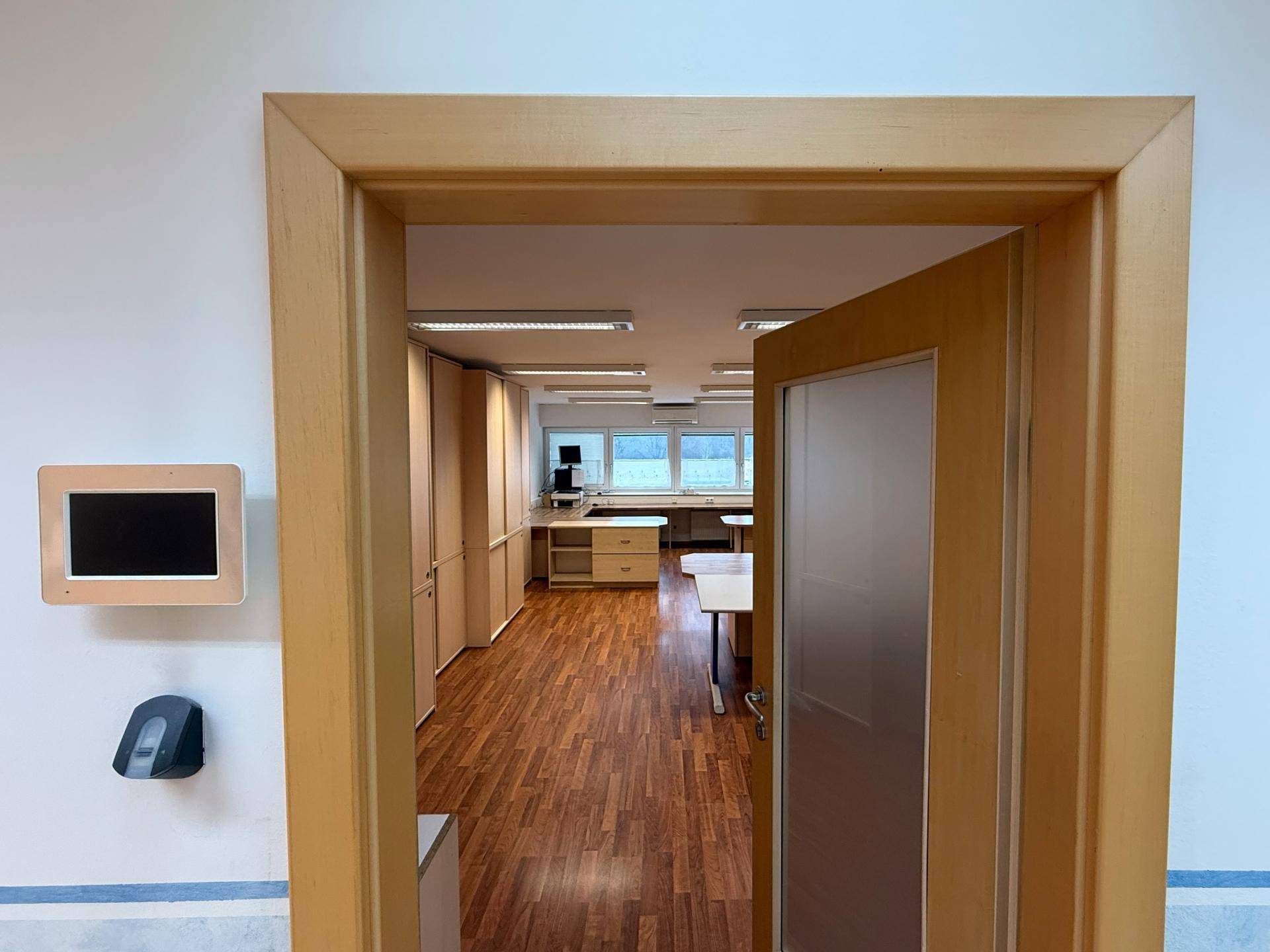 Offene Tür führt in ein helles Büro mit Holzmöbeln und großen Fenstern, einladender Eingangsbereich.