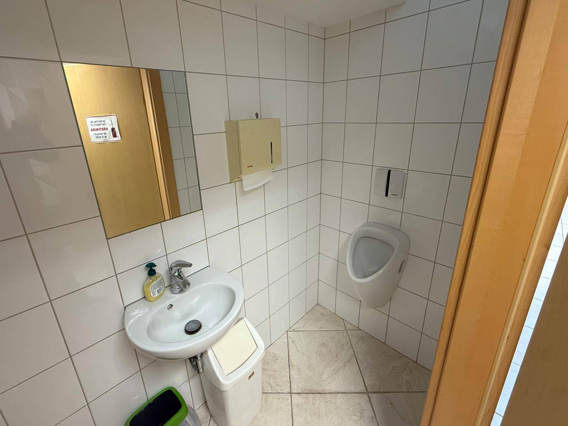 Kleiner, gefliester Herrenwaschraum mit Urinal, Waschbecken und Spiegel, sauber und funktional eingerichtet.