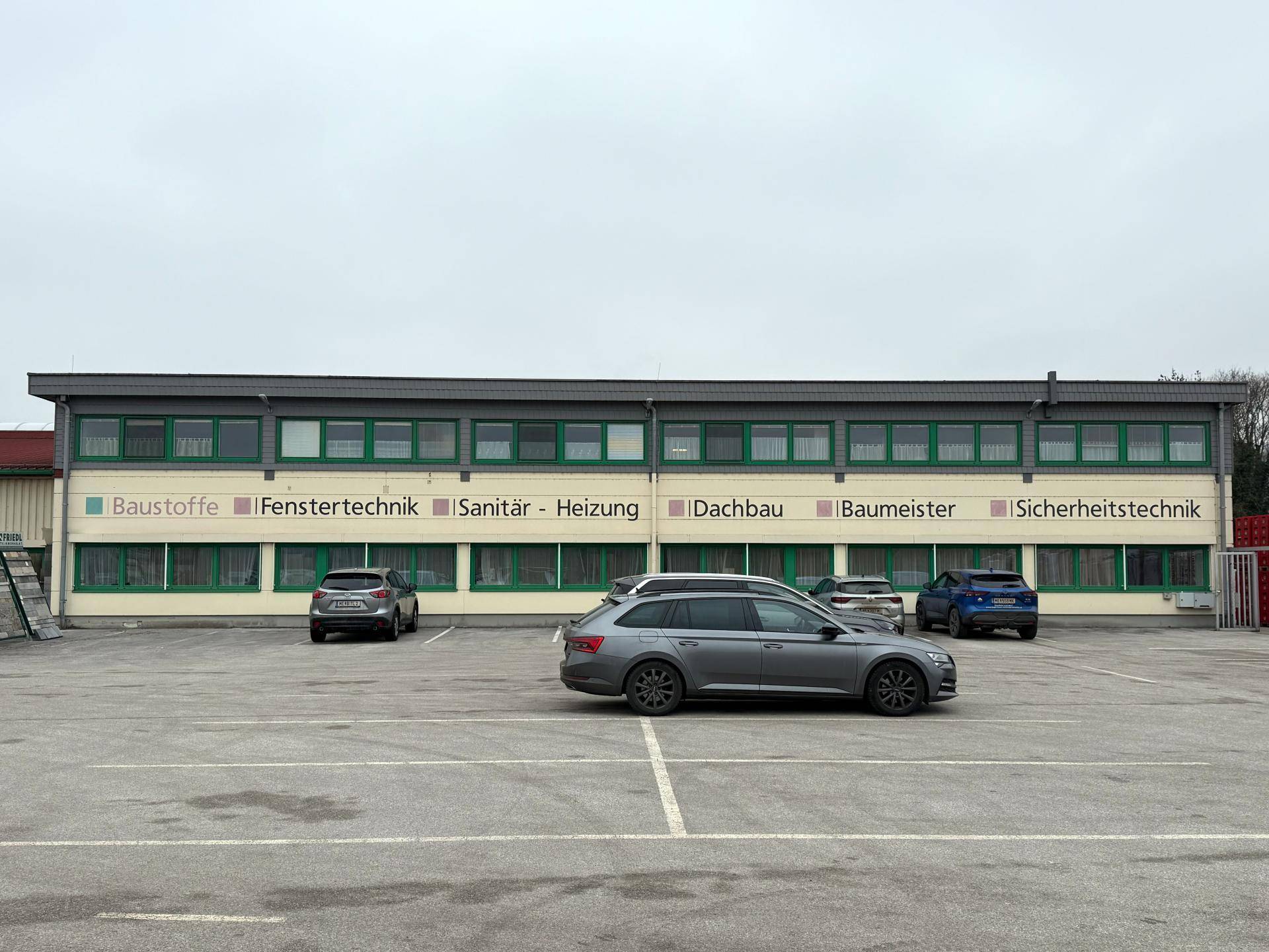 Moderne Außenansicht eines Geschäftsgebäudes mit grünen Fensterrahmen und einem weitläufigen Parkplatz davor.