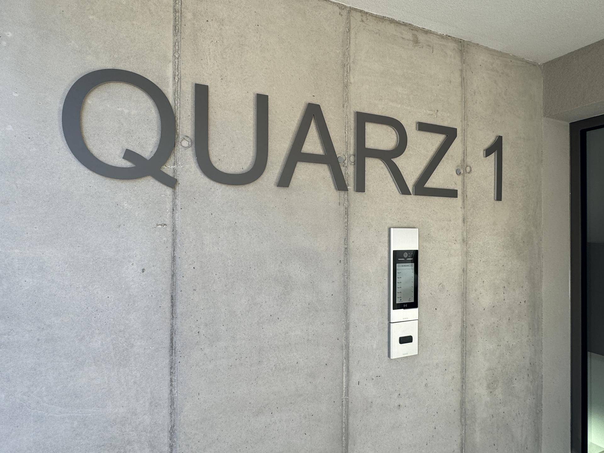 Moderner Eingangsbereich mit Betonwand, Gebäudename "QUARZ 1" und Gegensprechanlage.