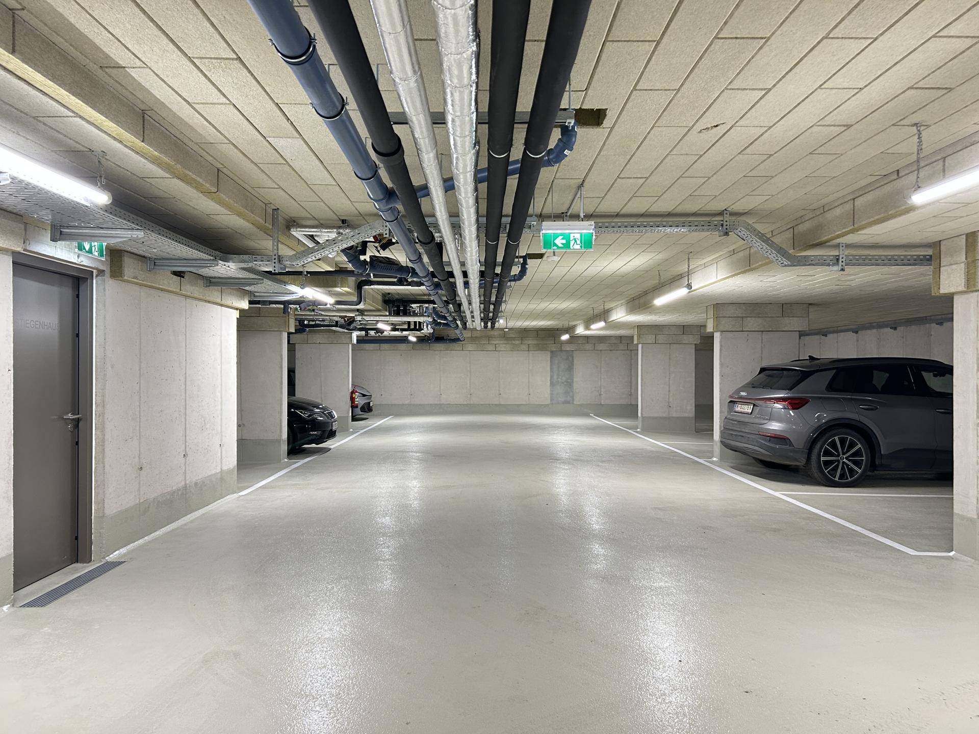 Geräumige Tiefgarage mit Betonwänden, sichtbaren Leitungen und markierten Parkplätzen.
